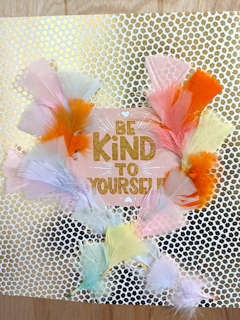Artwork with the words "Be Kind to Yourself" on a shiny metallic floral backing, adorned with colorful feathers