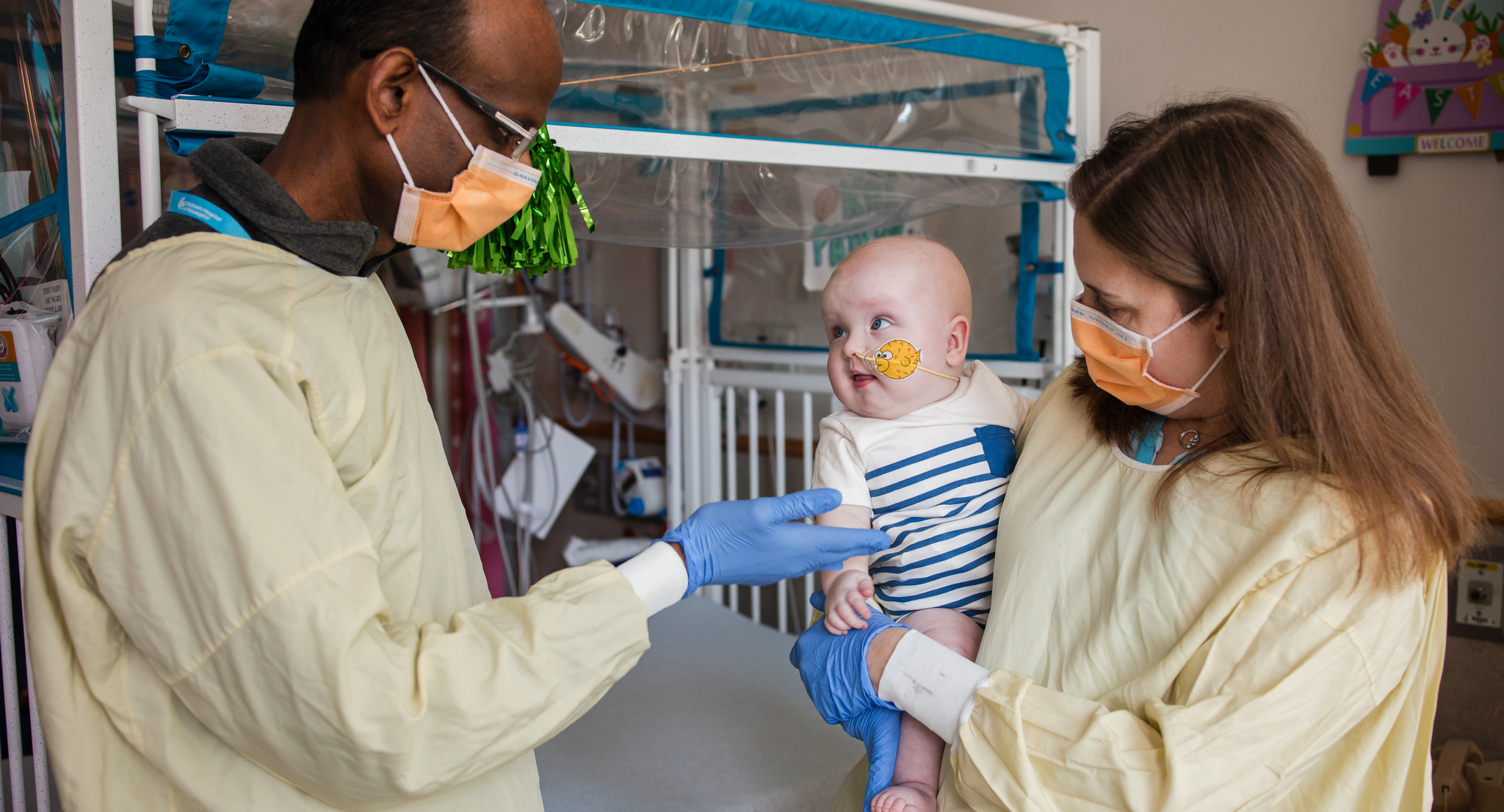 Rebecca Ahrens-Nicklas holds baby KJ while Kiran Musunuru gestures toward him; both adults masked and gowned