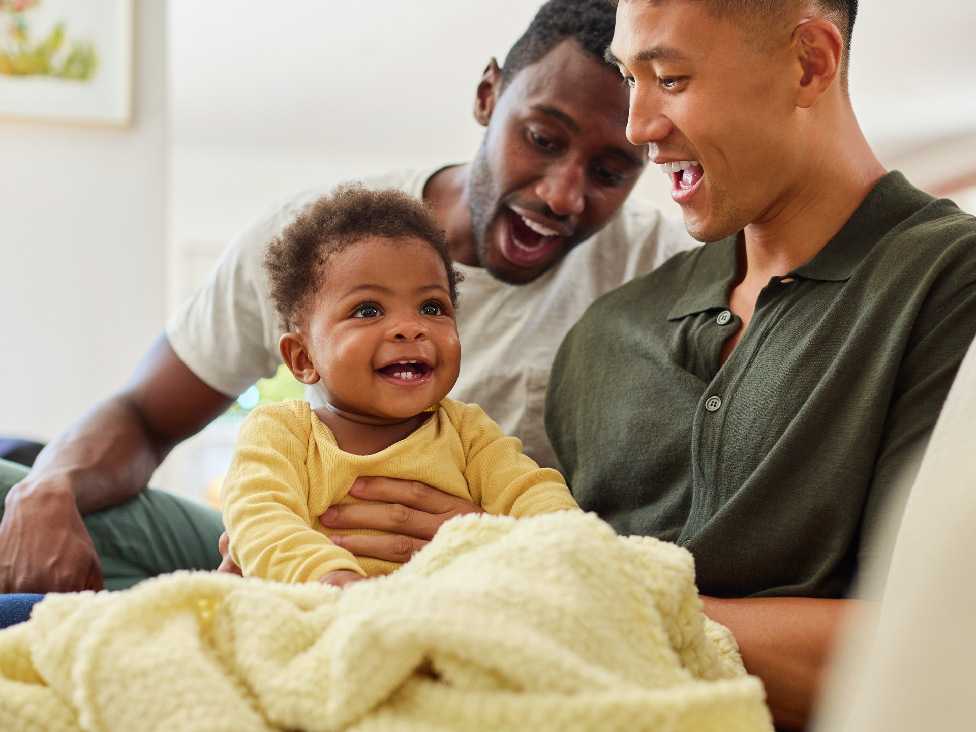 Same sex male couple or friends playing with baby sitting on sofa at home together