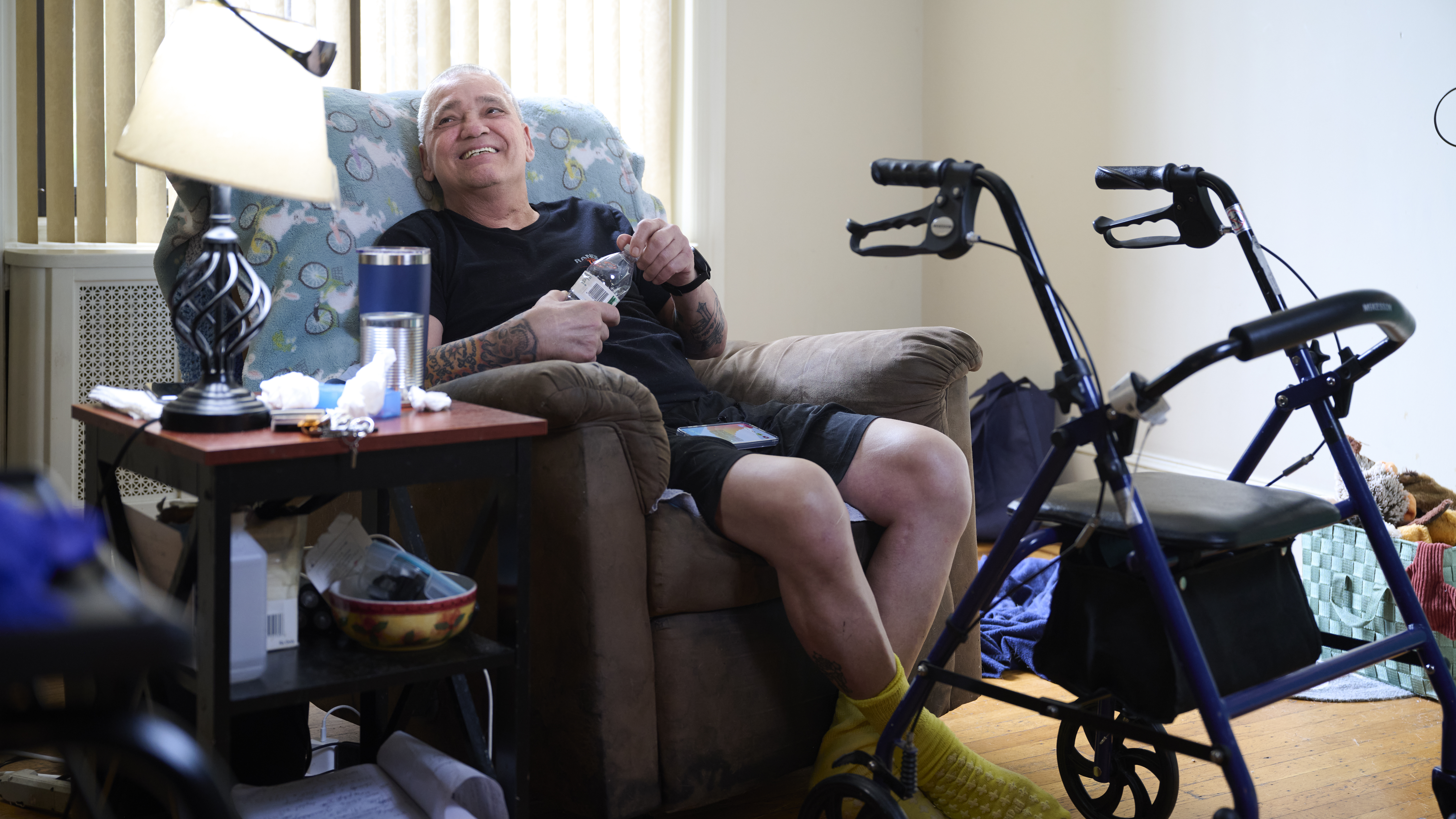Mike Desalis, a man in his early 60s, smiles and looks up while seated in a plush recliner chair surrounded by a walker and many personal items on a side table