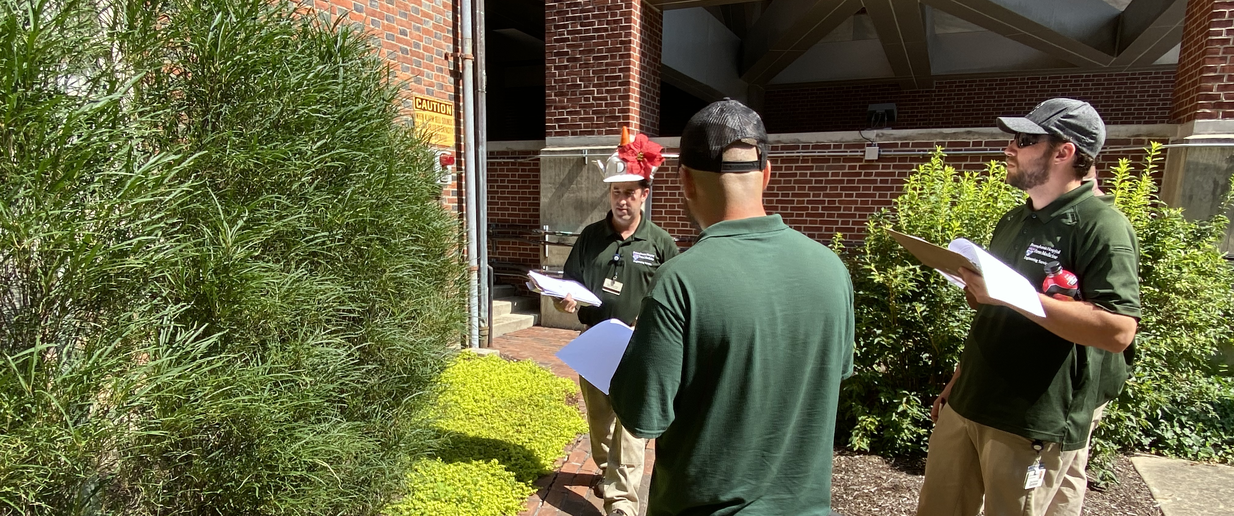 Dan Bangert stands in front of several plants in a courtyard. Nick Sambucetti and Blake Sieminski hold clipboards and examine the plants