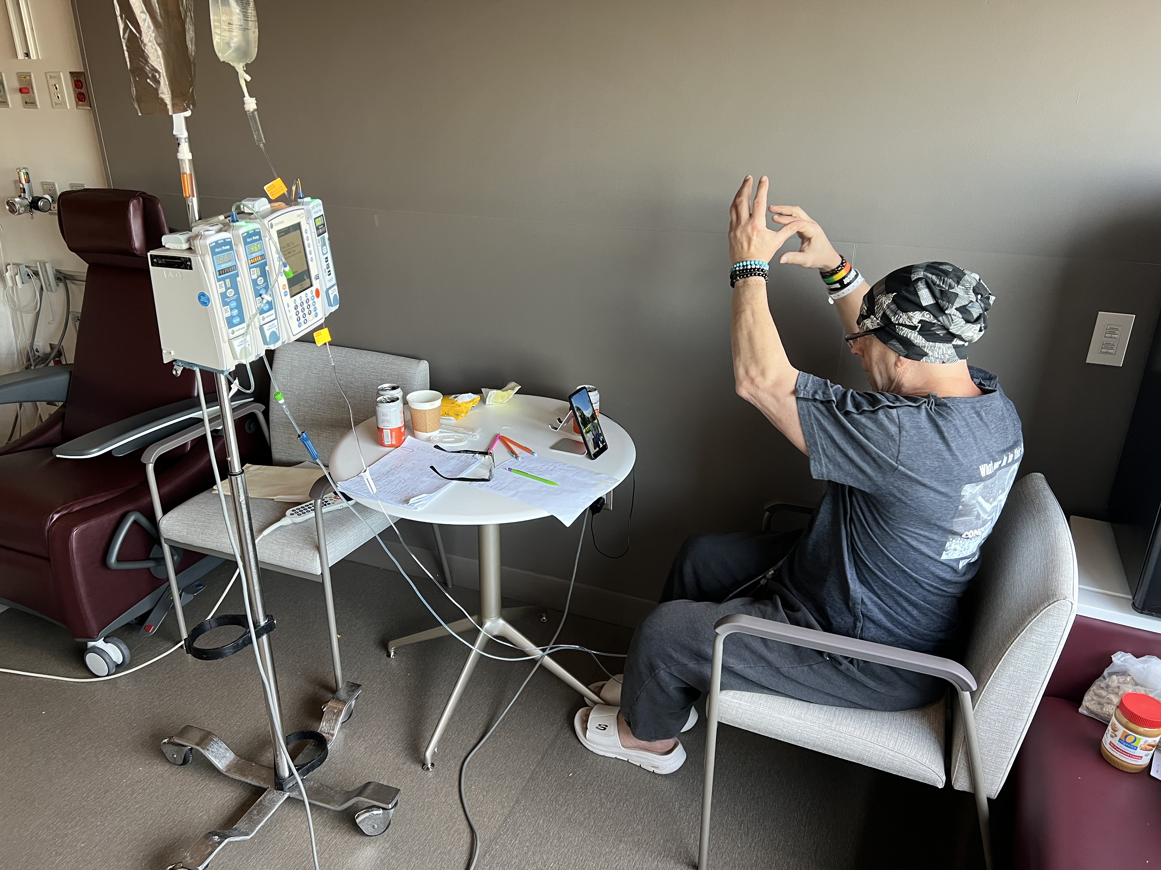 Howard Aaron sits in an armchair in his patient room, facing an iPhone with a video call, raising his arms above his head to instruct a client during a virtual workout.