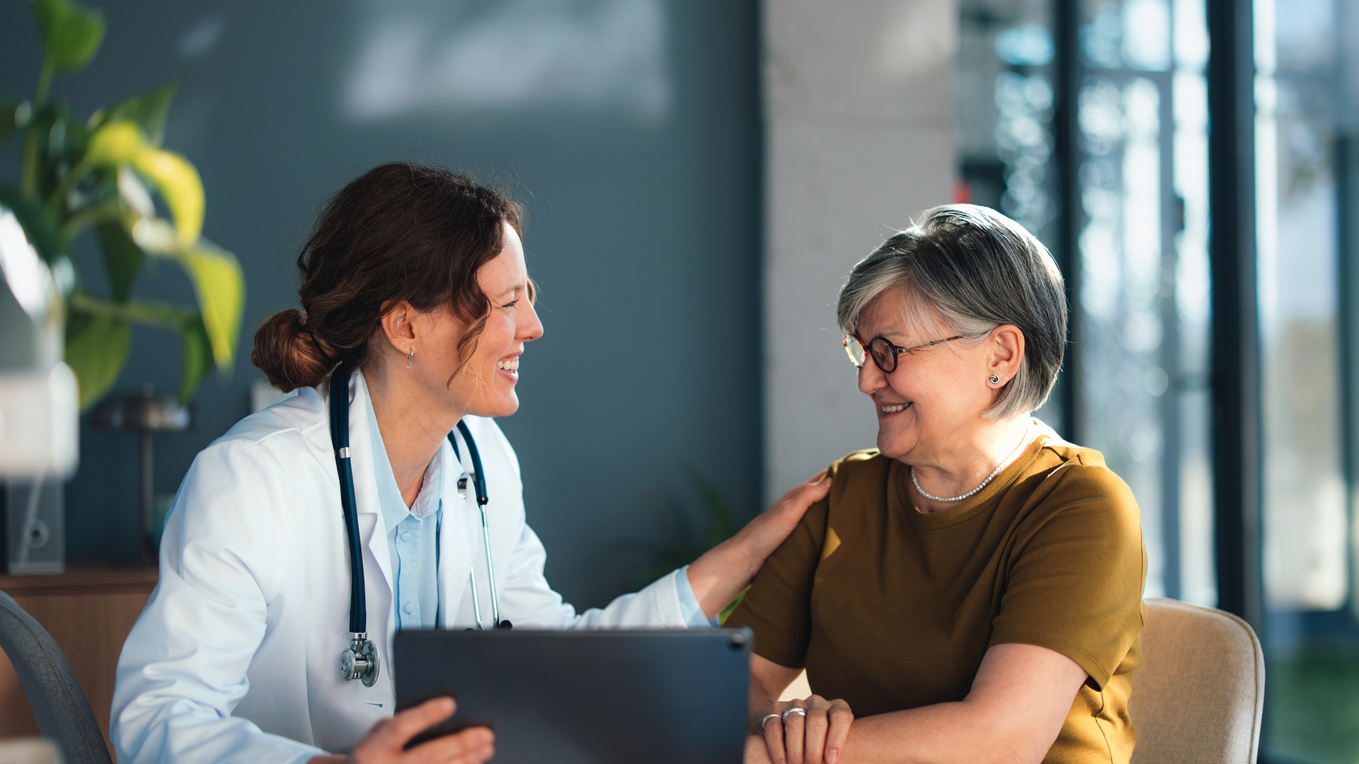 A female doctor gently consults a smiling senior woman in a bright clinic office, using a tablet while demonstrating trust, compassion and clear communication.