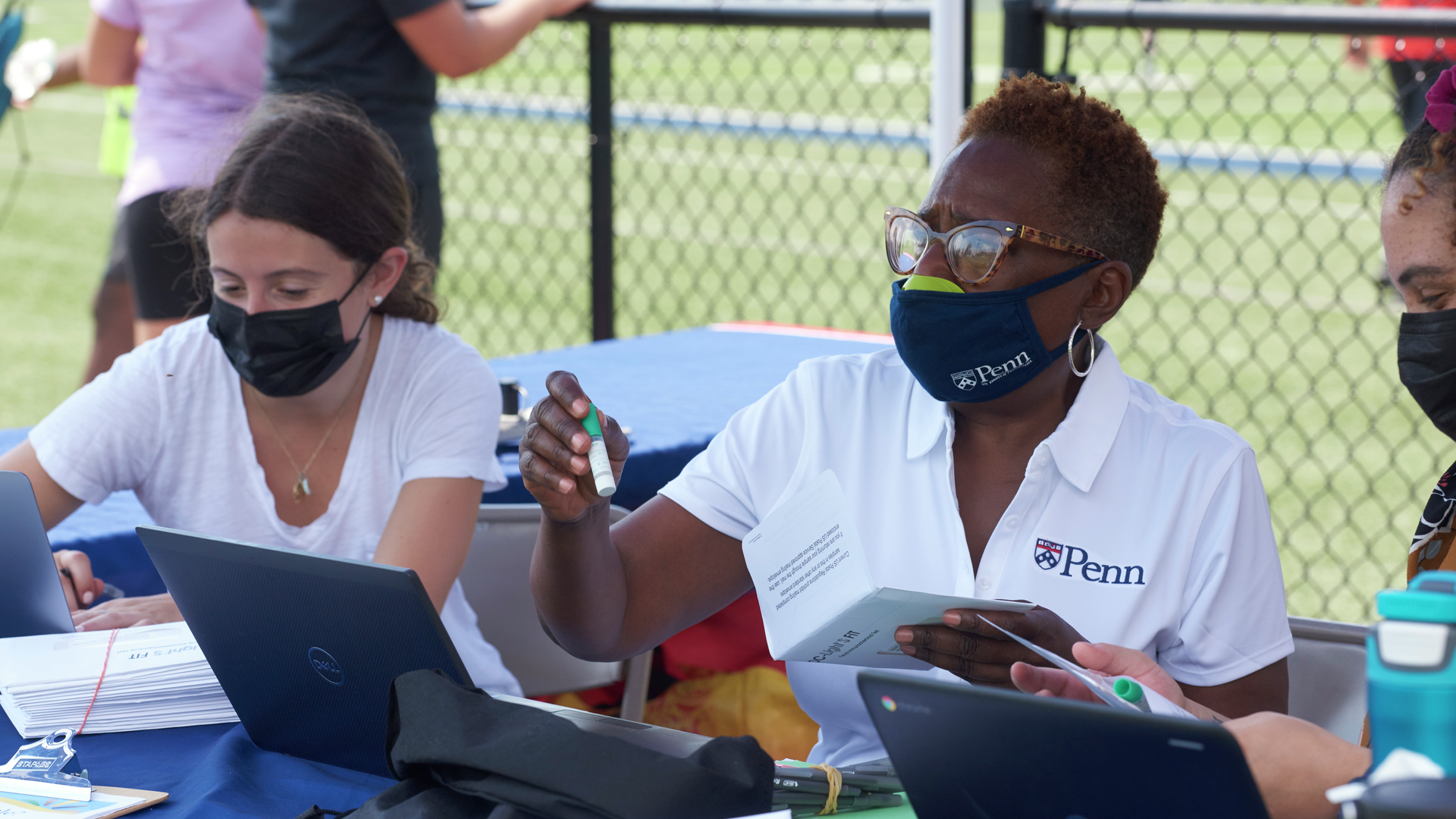 Claudia Melendez and Armenta Washington sit at an outdoor table and hand out FIT testing kits in envelopes