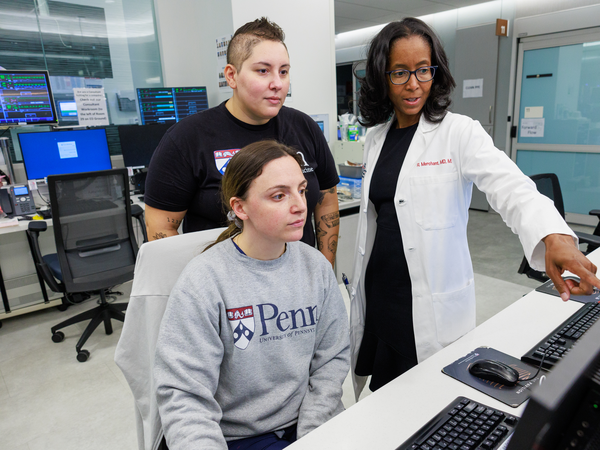 Raina Merchant, wearing a white coat, points at a computer screen with Emergency Department staff 