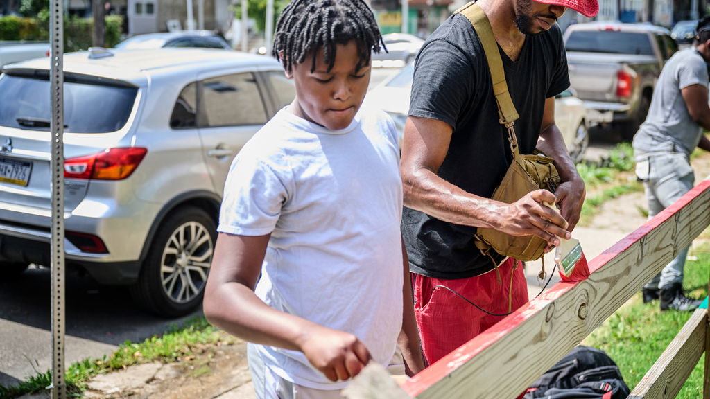 A teenage boy and a young man paint a fence around the periphery of a vacant urban lot