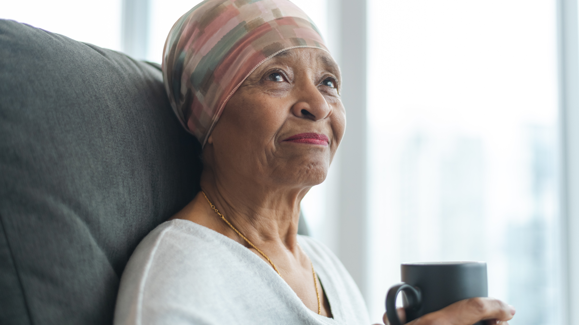 A black senior woman with cancer, wearing a scarf on her head, sitting in a chair looking contemplative