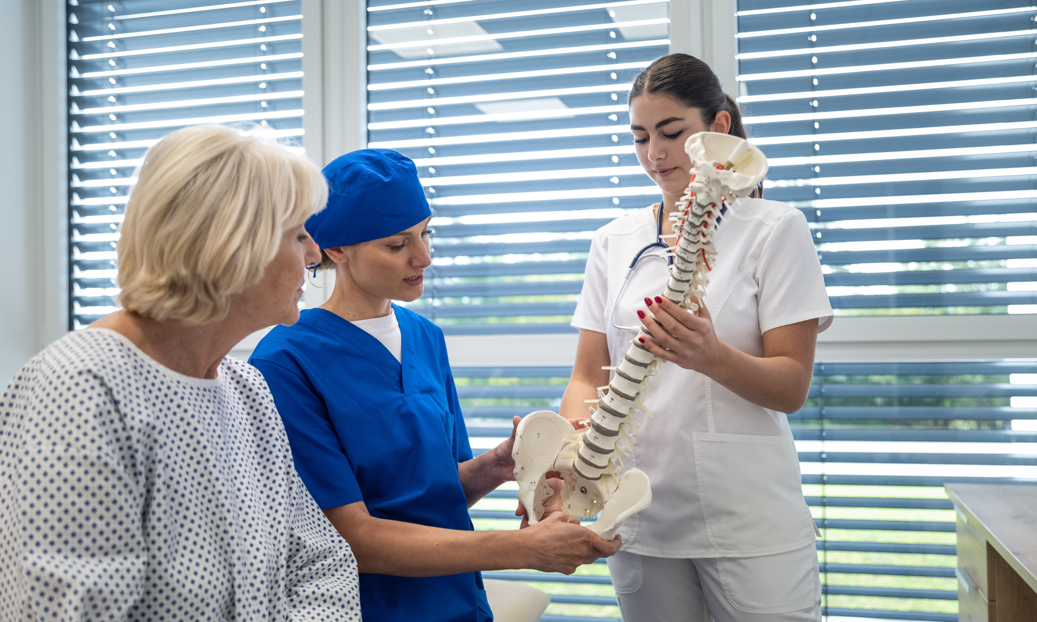A female surgeon in a blue scrub medical suit and a female doctor in a white uniform are talking to an elderly patient about the surgery