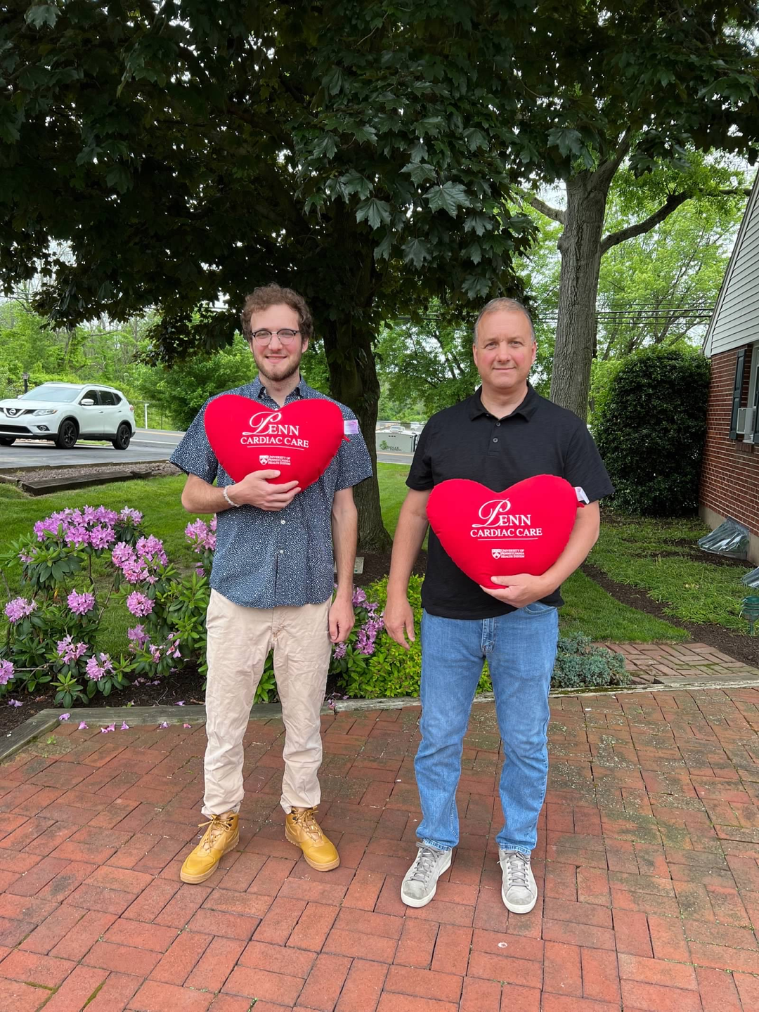 Ed and Tyler Schumacher stand on a suburban patio holding plush hearts labeled Penn Cardiac Care