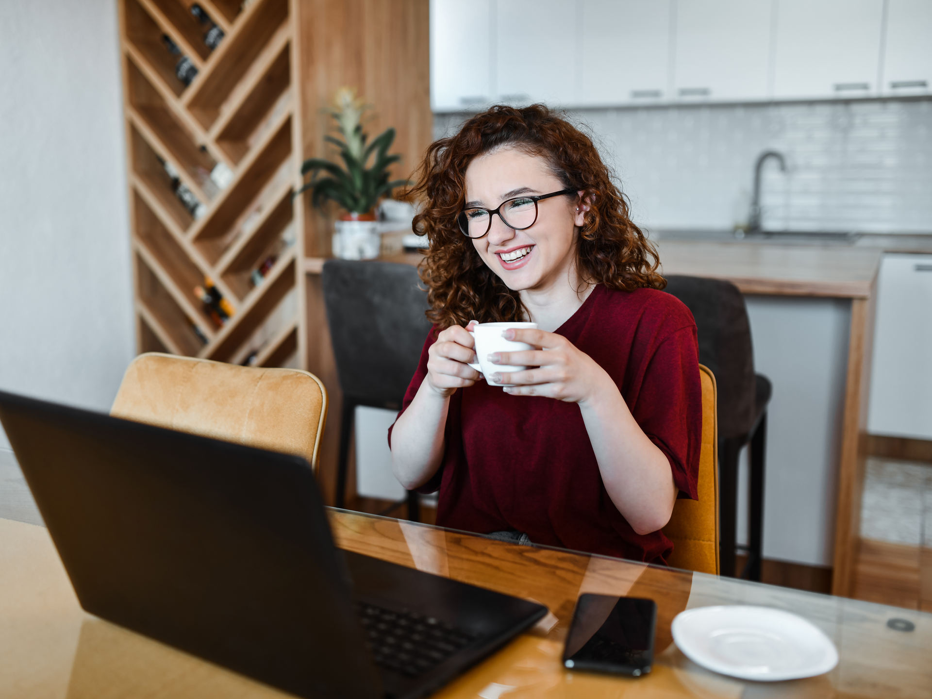 A young woman implied to be on a video call at her dining room table holding a mug, smiling at her computer.