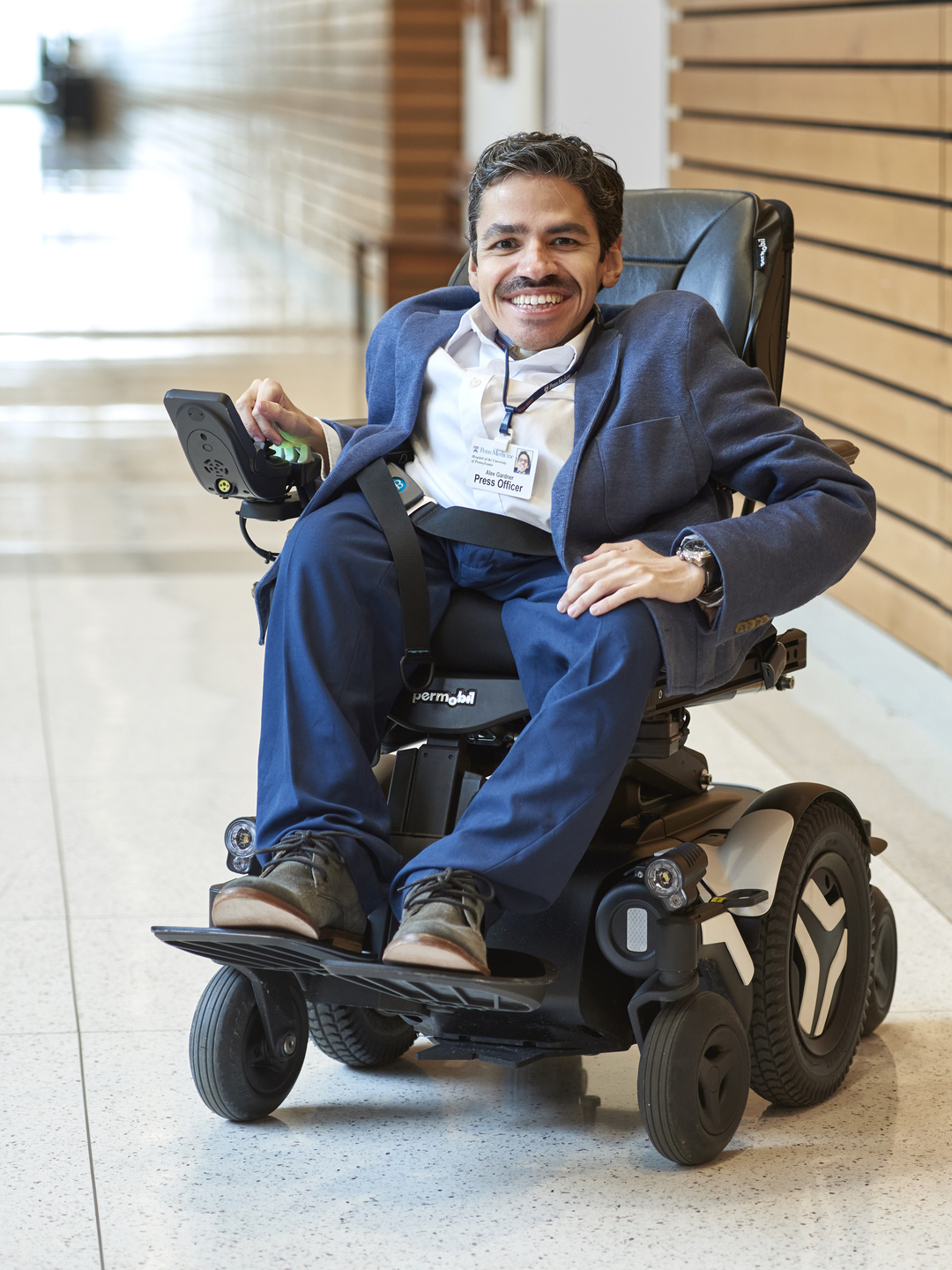 Alex Gardner, seen in a full body photo sitting in a wheelchair, relaxed and smiling, with his right hand on the chair's control panel