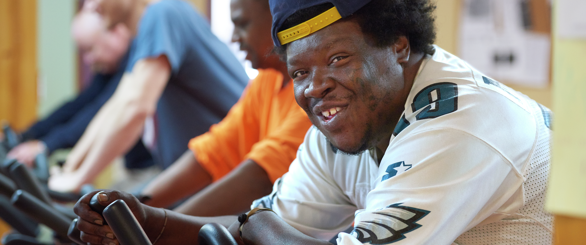 A man wearing a backwards cap and an Eagles jersey gives a knowing gap-toothed smile while riding an exercise bike