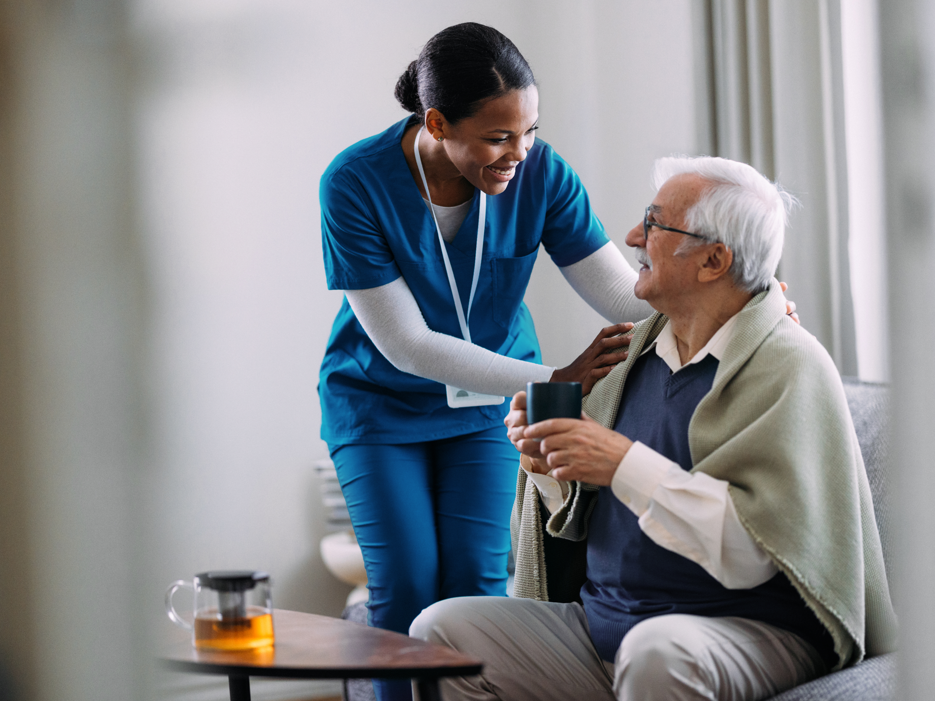 A compassionate nurse in a blue uniform helps an elderly man with a drink, creating a warm and caring atmosphere in a home setting. The scene highlights caregiving and support.