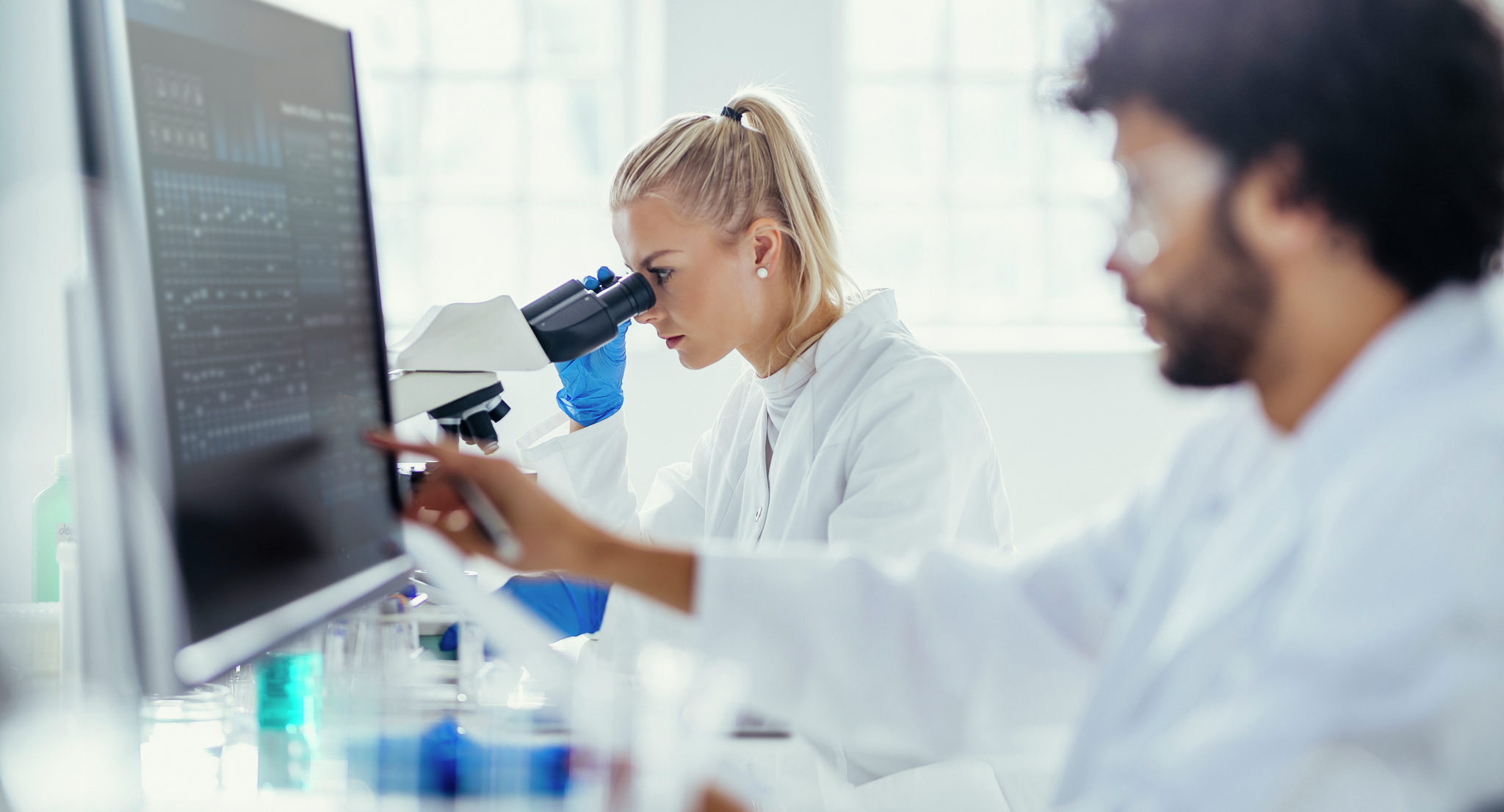 Two researchers concentrate on analyzing specimen under a microscope, amidst scientific tools in a bright laboratory setting.