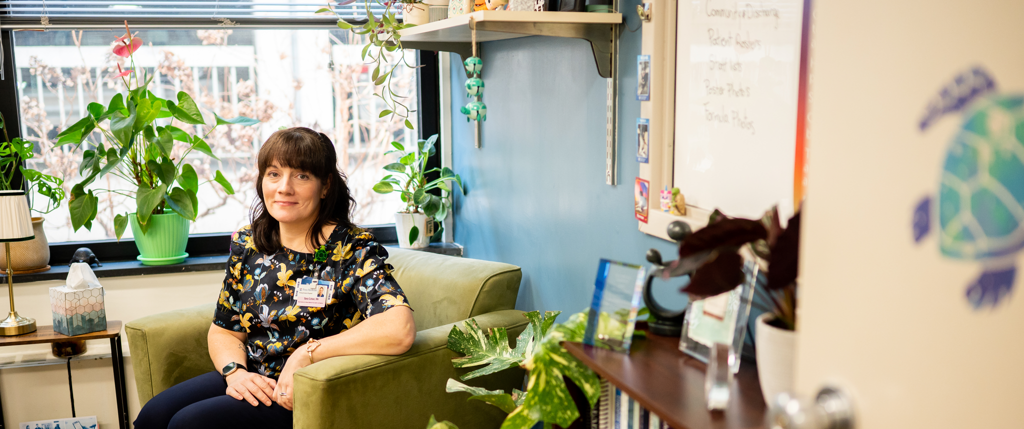 Sara Cohen sitting in a chair by a window in her office, surrounded by potted plants