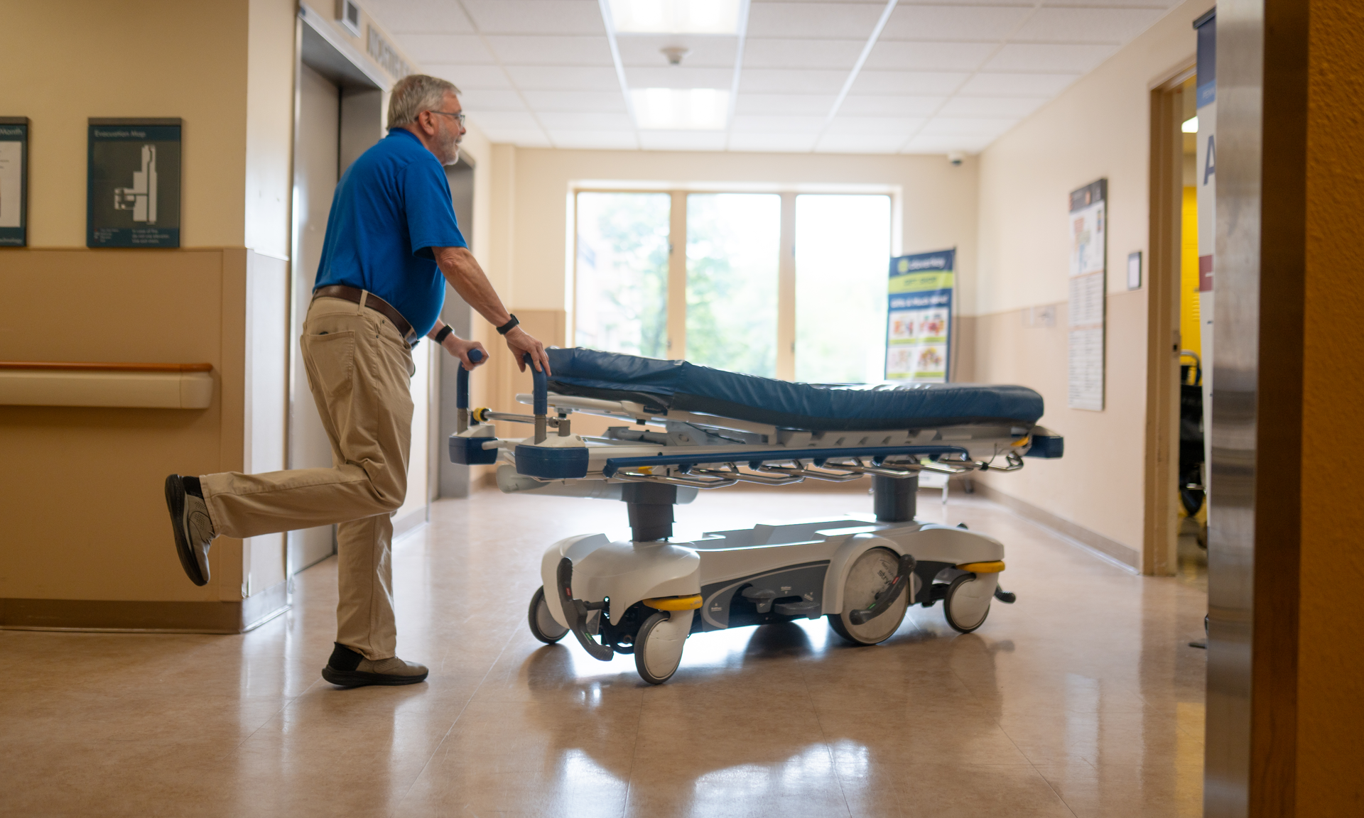 Ed McIntyre, patient transport volunteer, is pictured pushing a patient transport bed through the hospital hallway. The hallway is illuminated in light coming from the window, and Ed has his one foot propped in the air as he pushes the cart