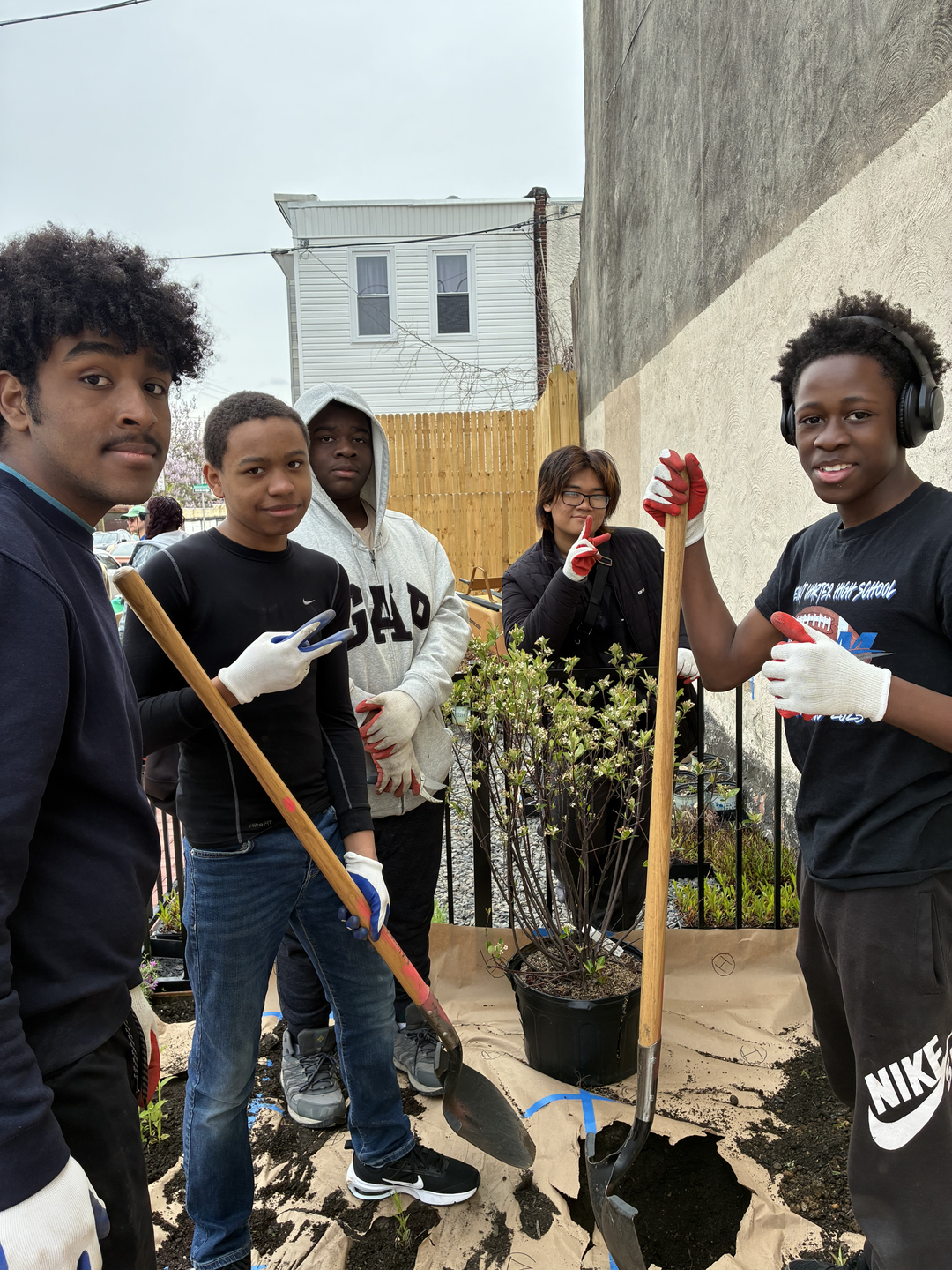 Five young people in gardening gloves use tools to dig a hole for a plant in a pot