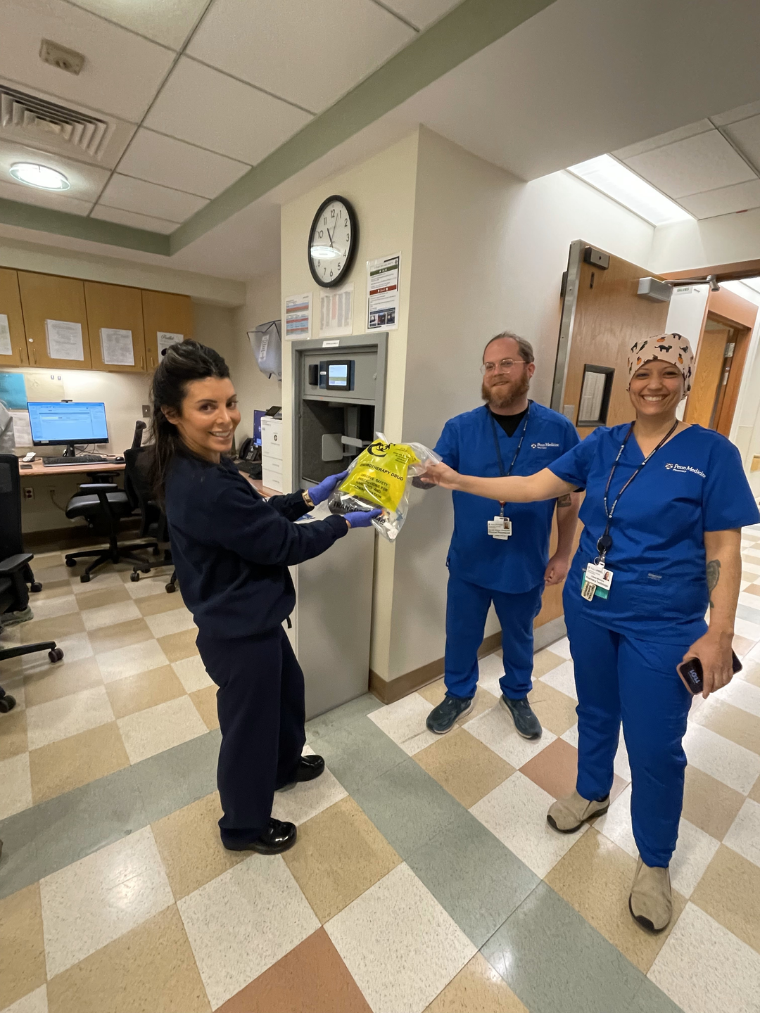 Two clinical team members in scrubs hand a bag to a colleague in a hospital hallway