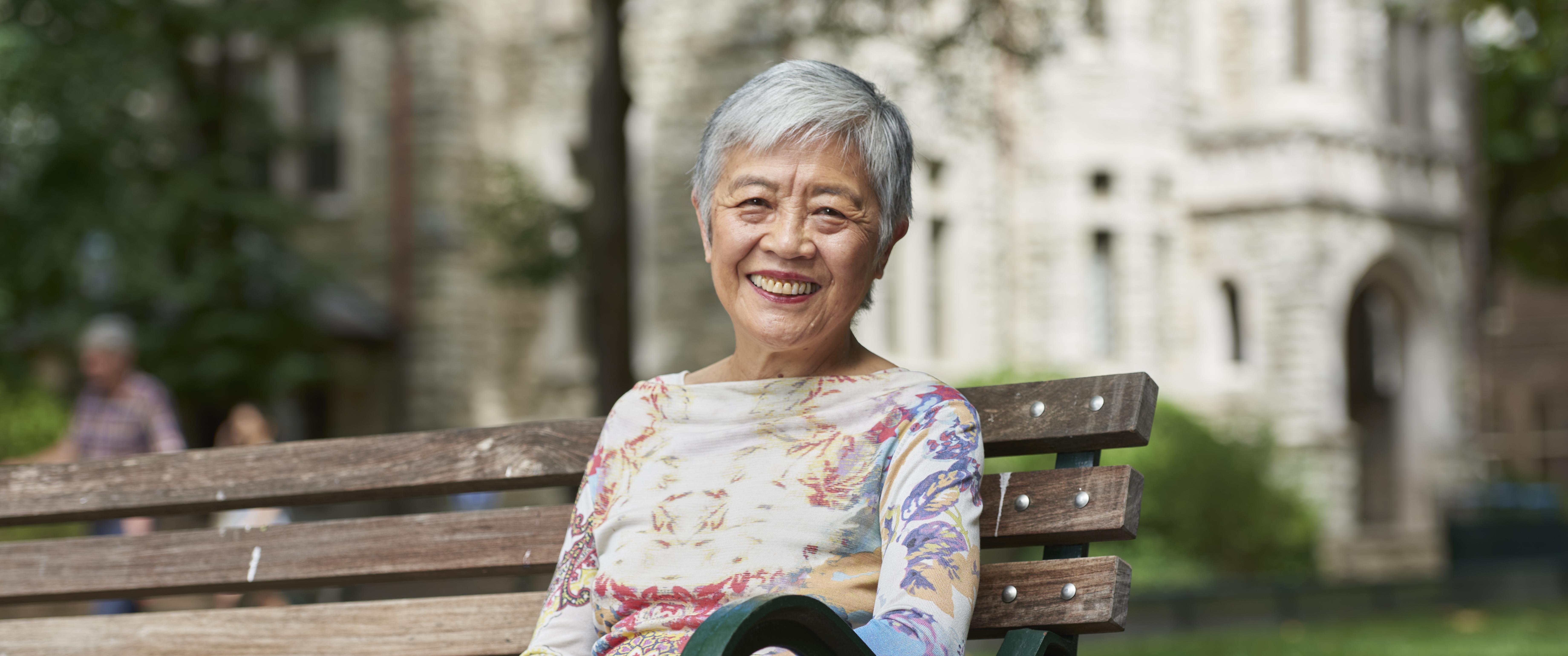 Portrait of Virginia M.-Y. Lee sitting on a campus bench, smiling, wearing colorful clothing