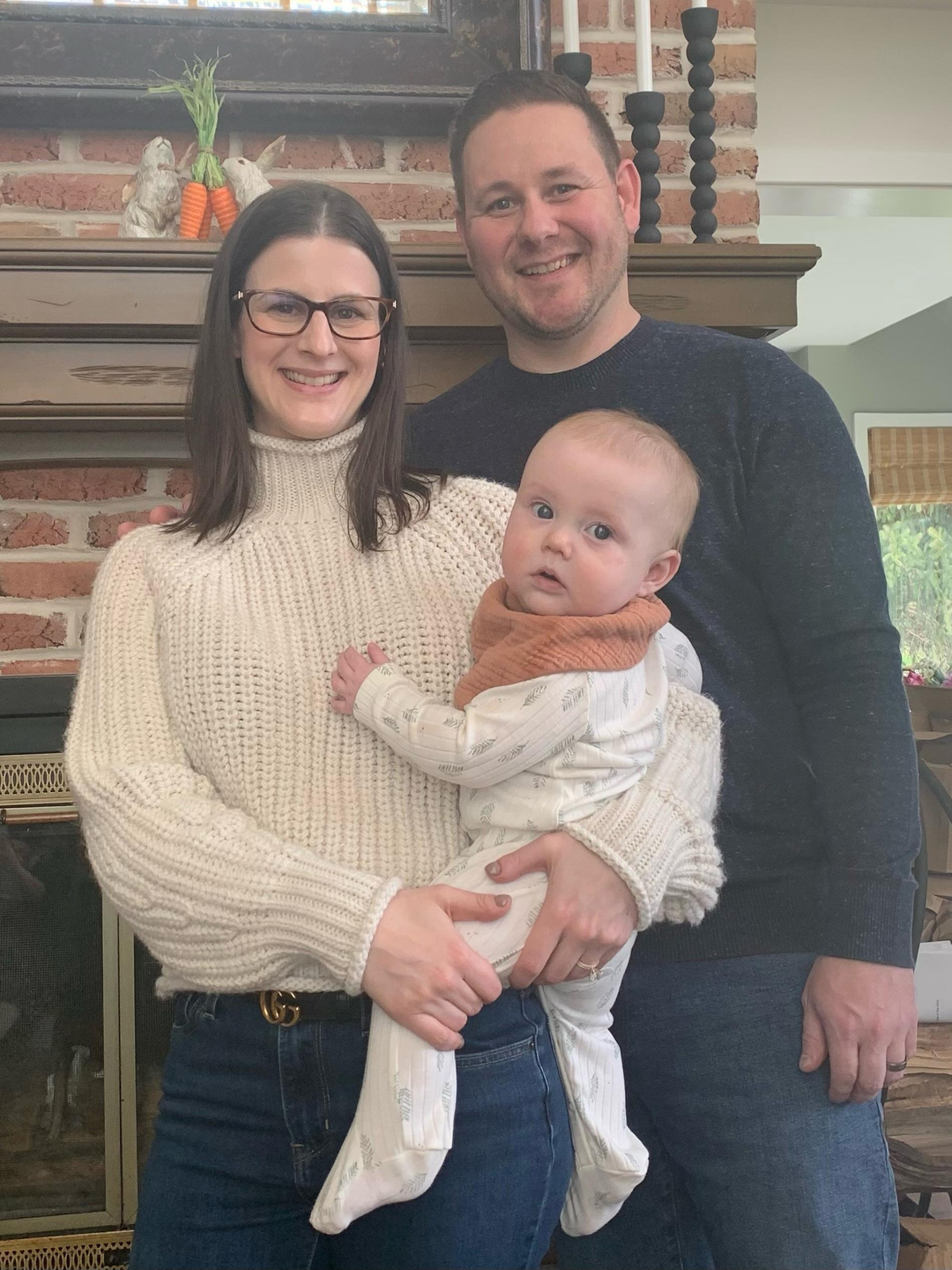 Carly Albright Keller, holding baby Rory in her arms, stands next to her husband Justin while they all smile and pose for a photo in their home.
