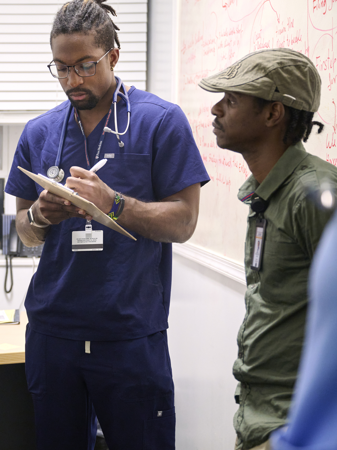 A person in scrubs takes notes on a clipboard, surrounded by other staff and volunteers