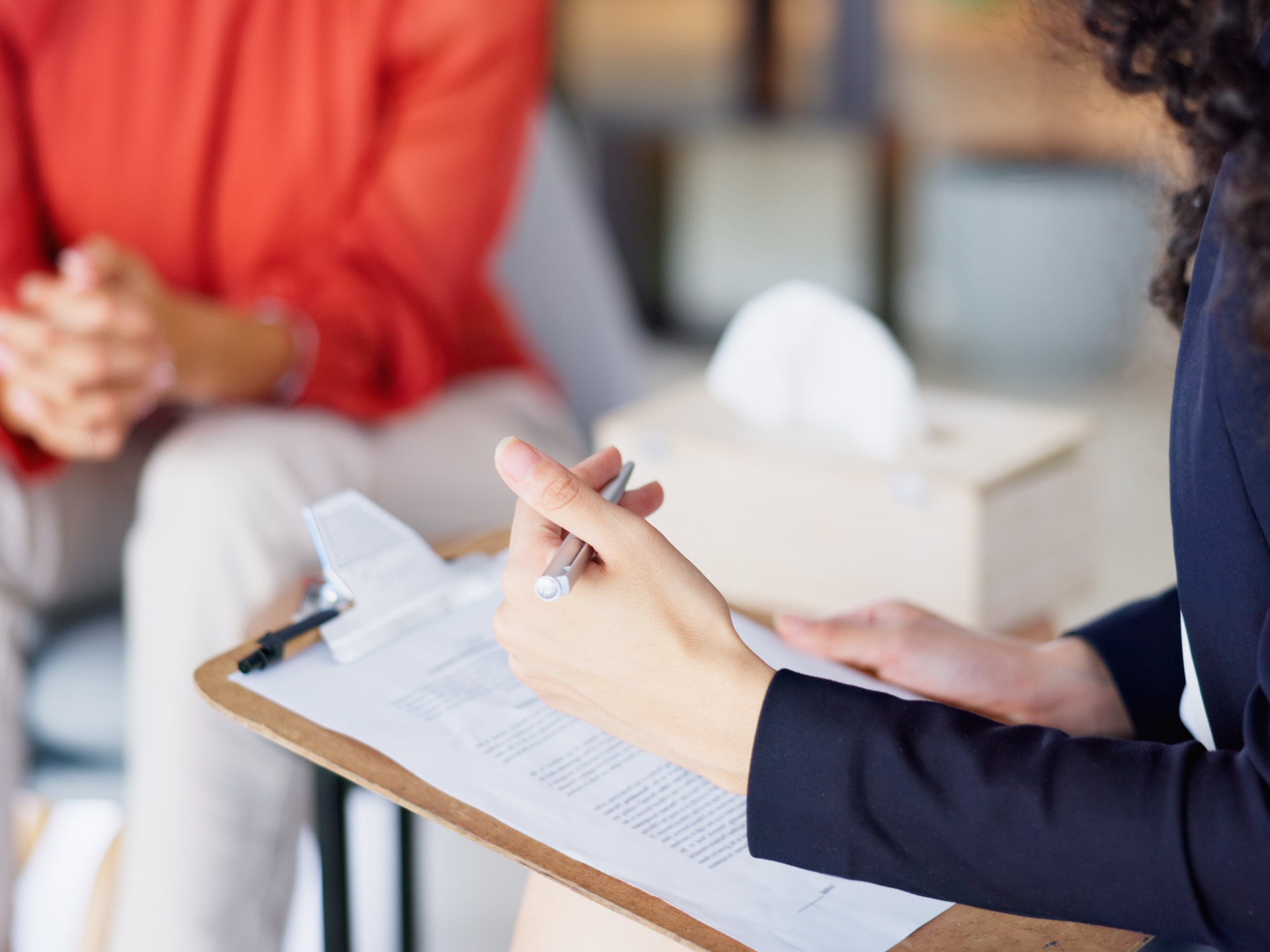 A therapist in a blue blazer writing notes down on a clipboard while sitting across from her patient.