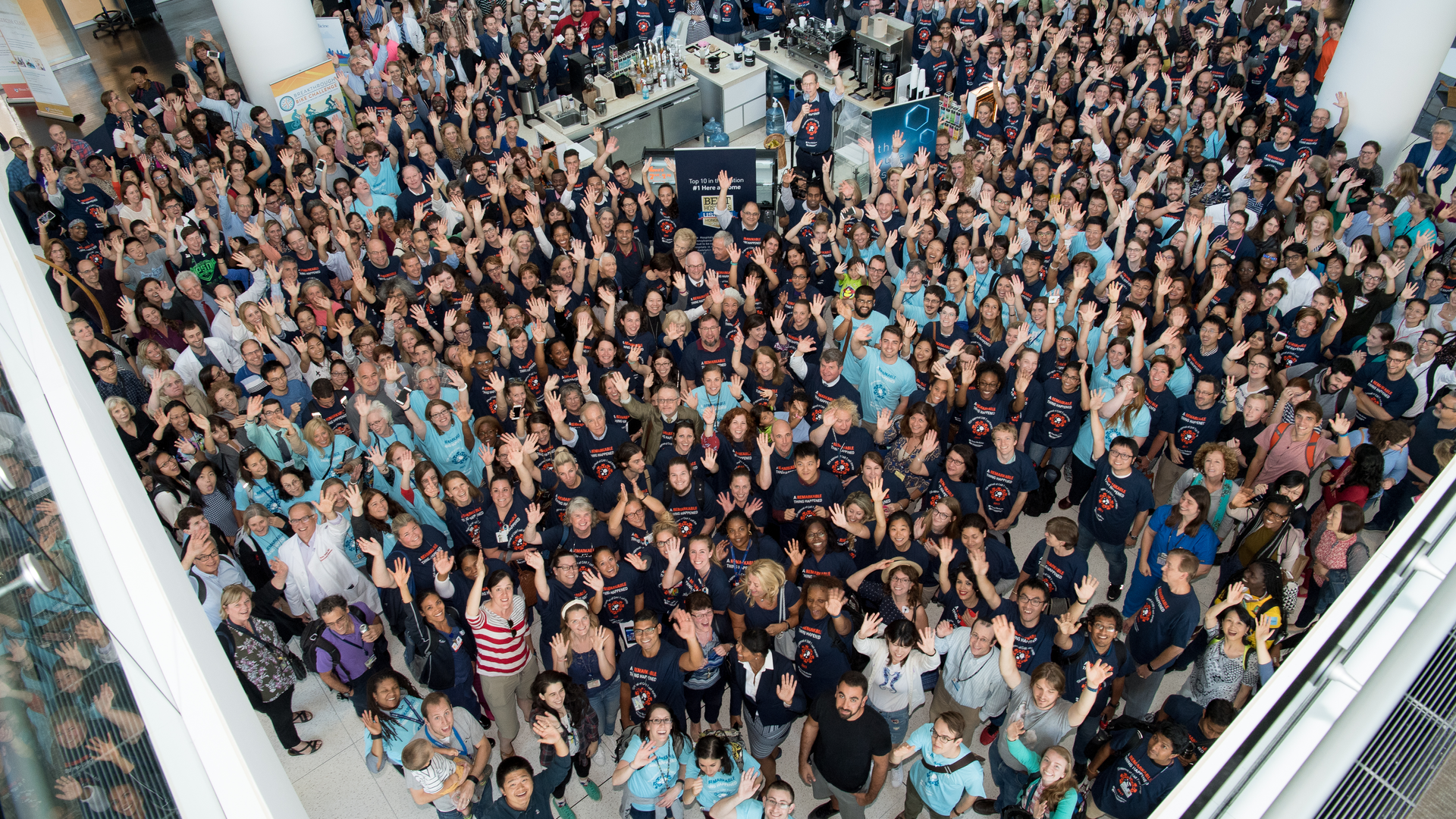 A crowd of hundreds of people, many wearing matching dark blue or light teal T-shirts, seen from above smiling and waving, fill all the space of the first-floor atrium of the Perelman Center for Advanced Medicine