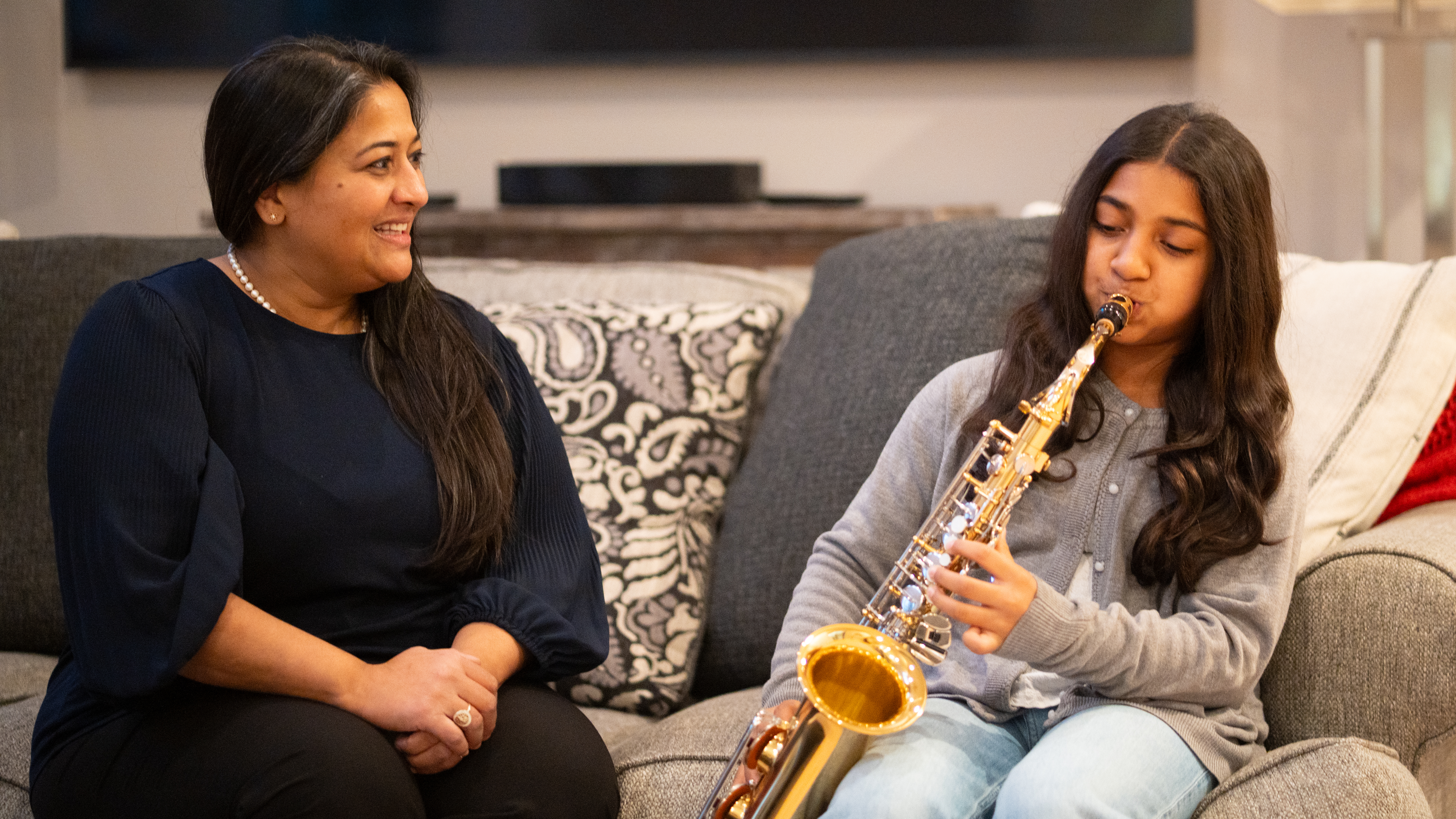Aileen John and her daughter Madison sit on the couch while Madison practices the saxophone