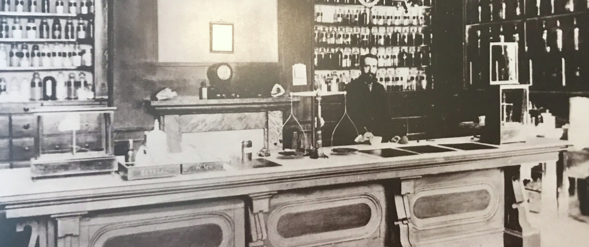 An old black and white photo of a large stone Apothecary desk. A bearded man stands behind it, and behind him is a wall with small drawers with shelves above in arched alcoves, filled with labeled jars and bottles