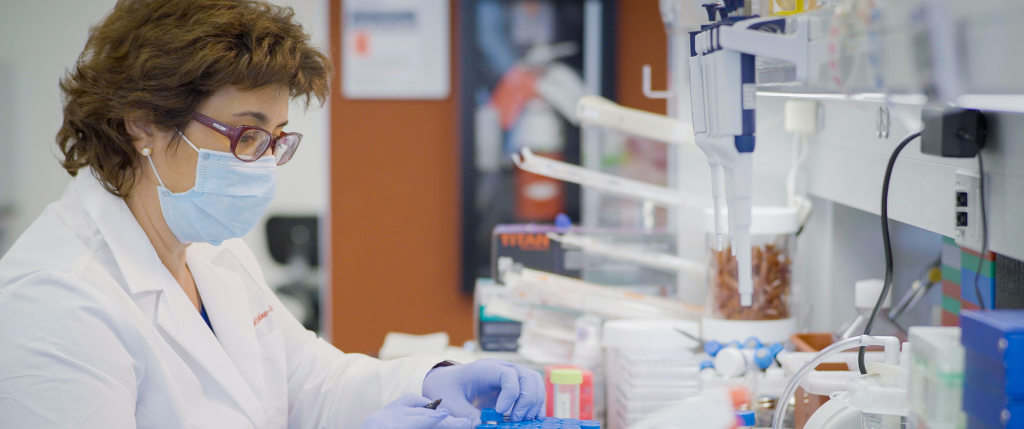 A woman wearing a lab coat, mask, and gloves, works at a lab bench doing mRNA research