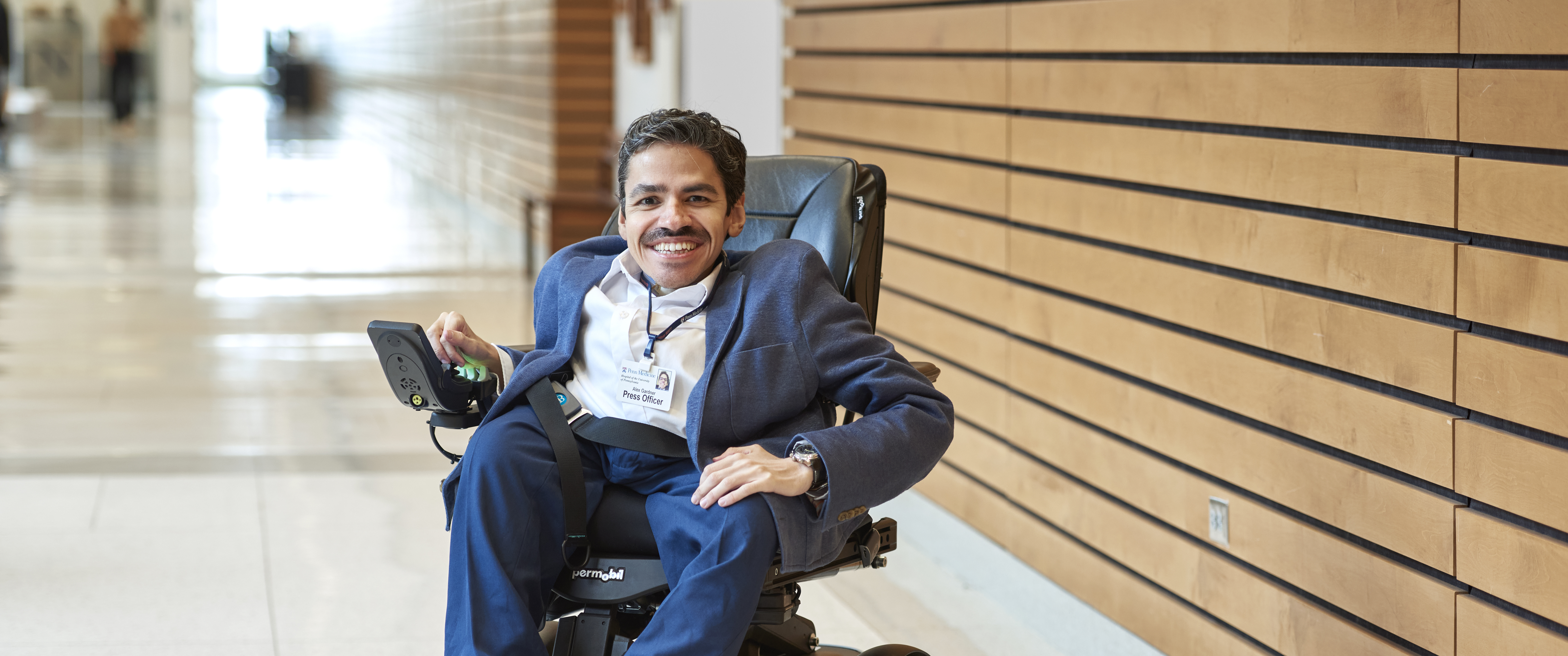 Alex Gardner, seen in a full body photo sitting in a wheelchair, relaxed and smiling, with his right hand on the chair's control panel