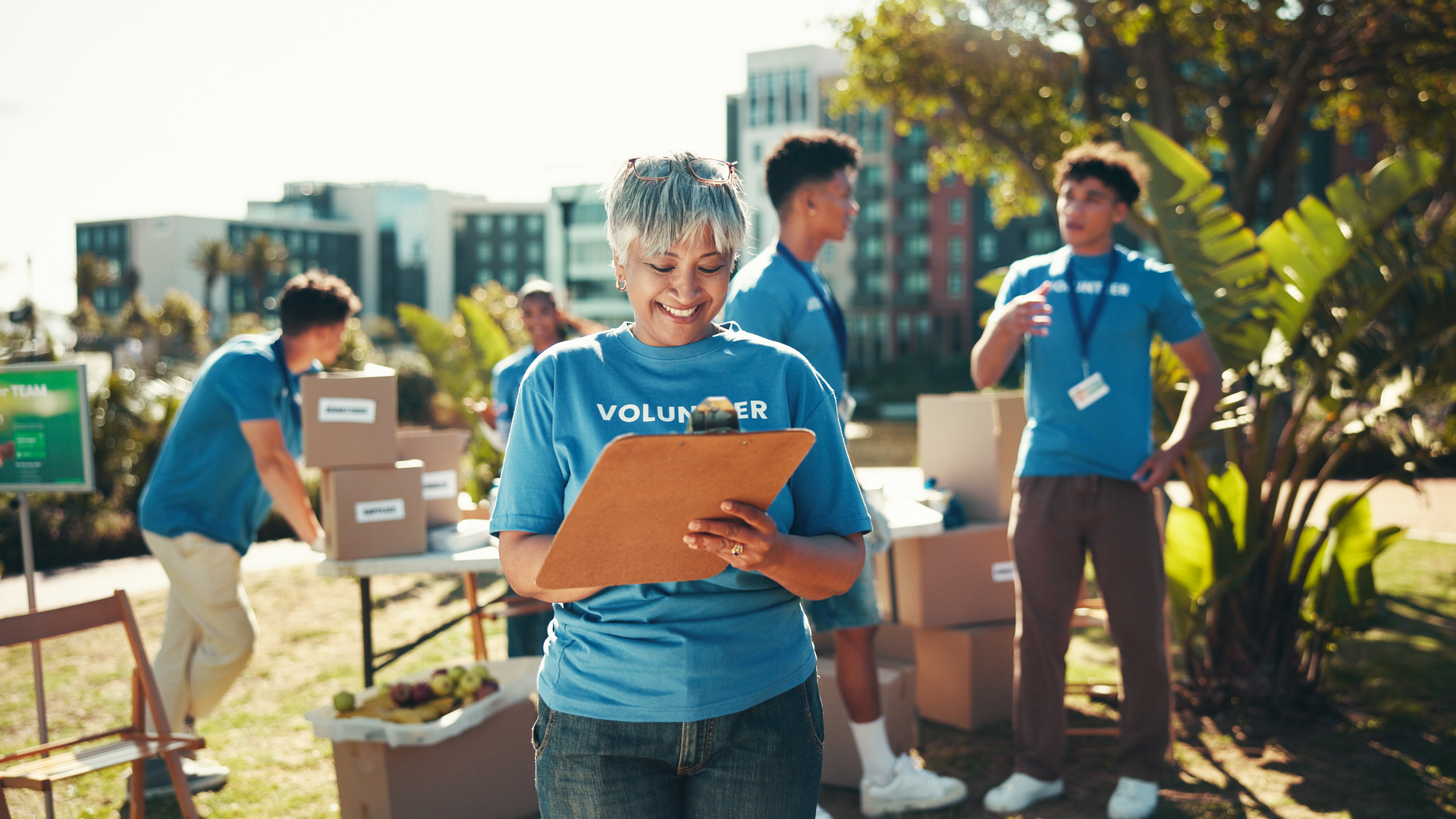 Happy, volunteers and mature woman with clipboard, charity and inventory for donations, smile and ngo. Outdoor, team and coordinator with checklist for info, community service and boxes for nonprofit