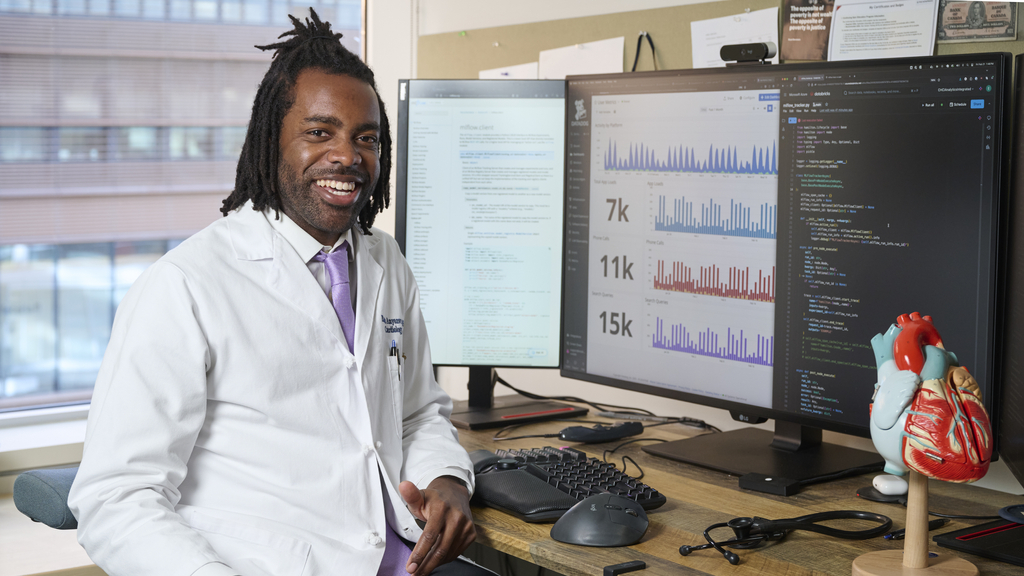 Emeka Anyanwu in a white coat, seated at a desk with data charts displayed on his monitor and a model heart nearby 