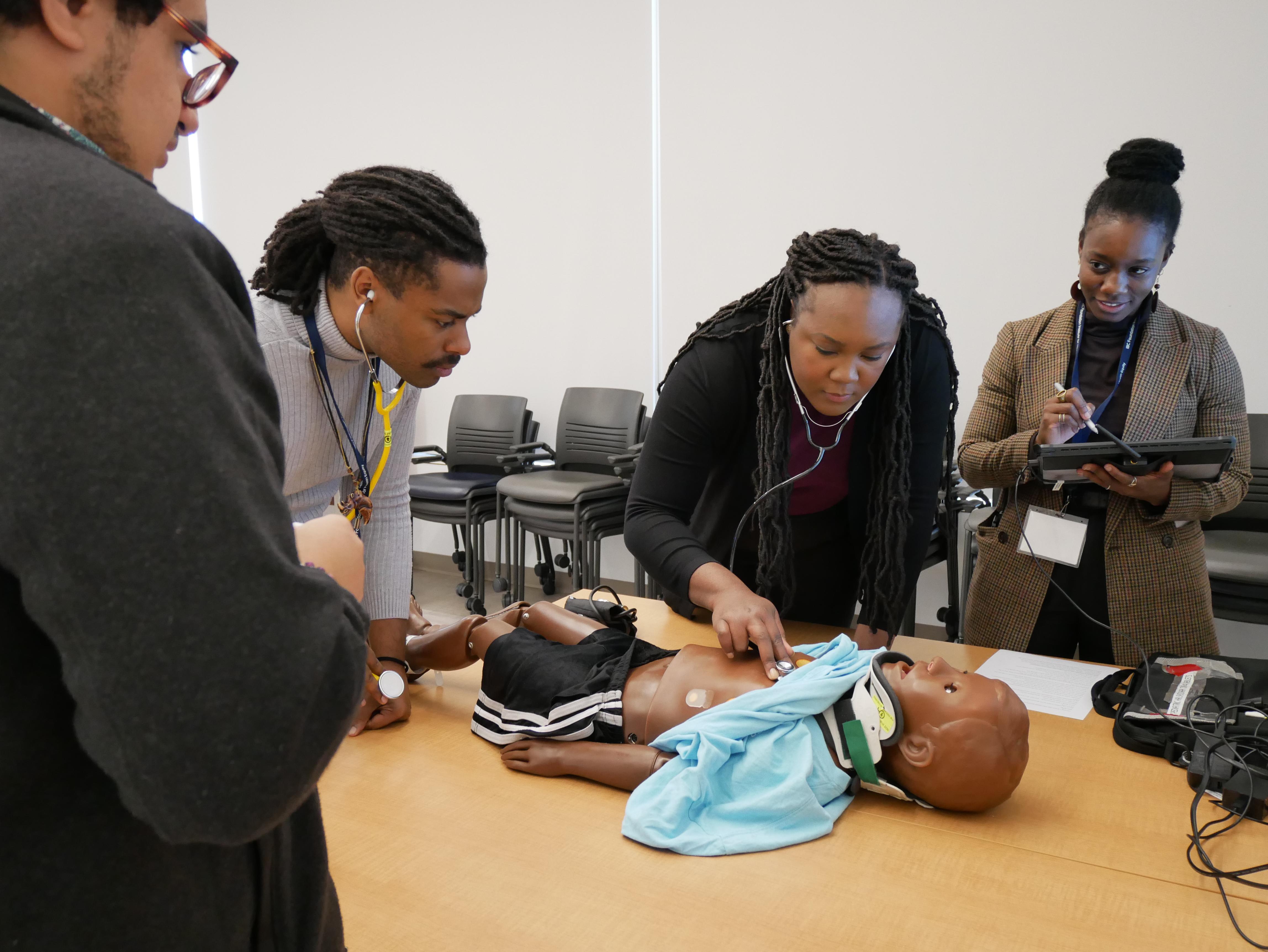 Claudia Gambrah-Lyles, MD, wearing a tweed blazer and holding a tablet, watches as three Black medical students monitor a child-sized manikin representing a trauma patient.