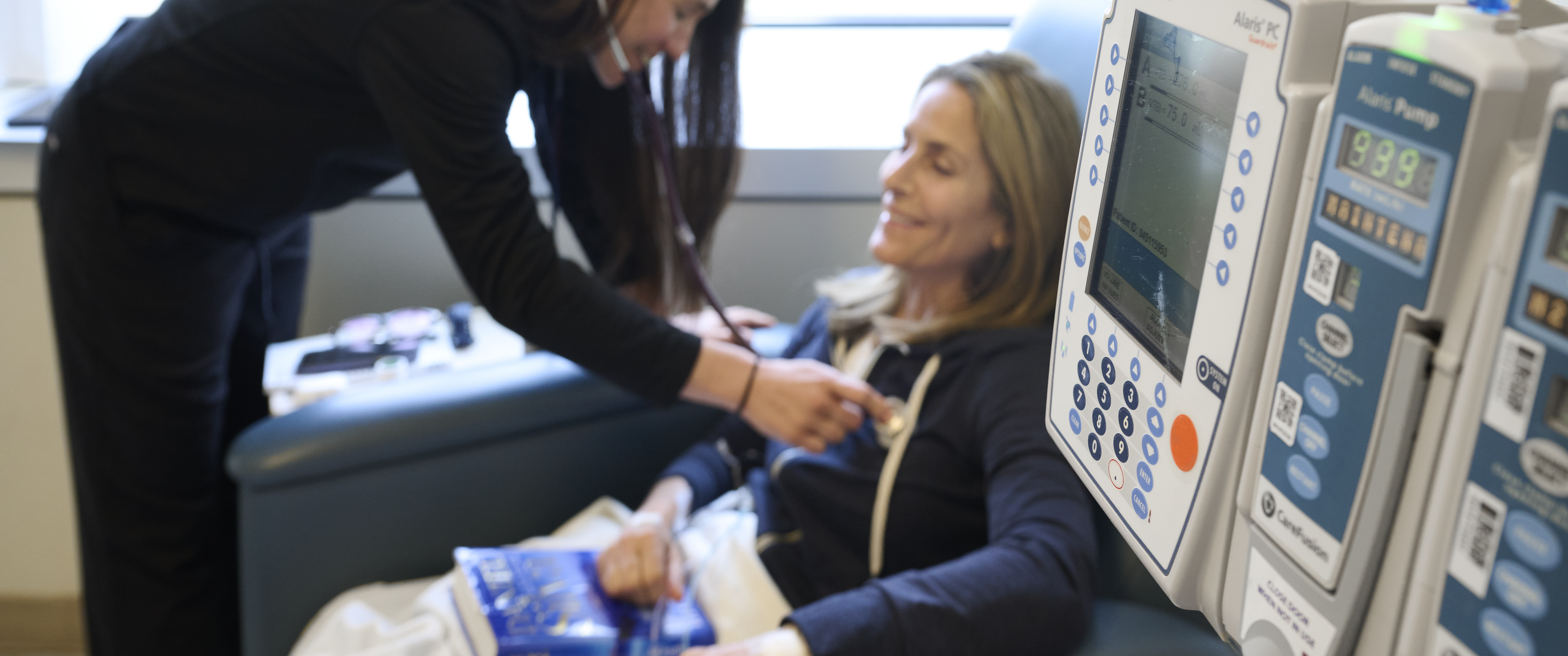 An infusion pump is in the foreground with an out-of-focus female patient in an infusion chair in the background
