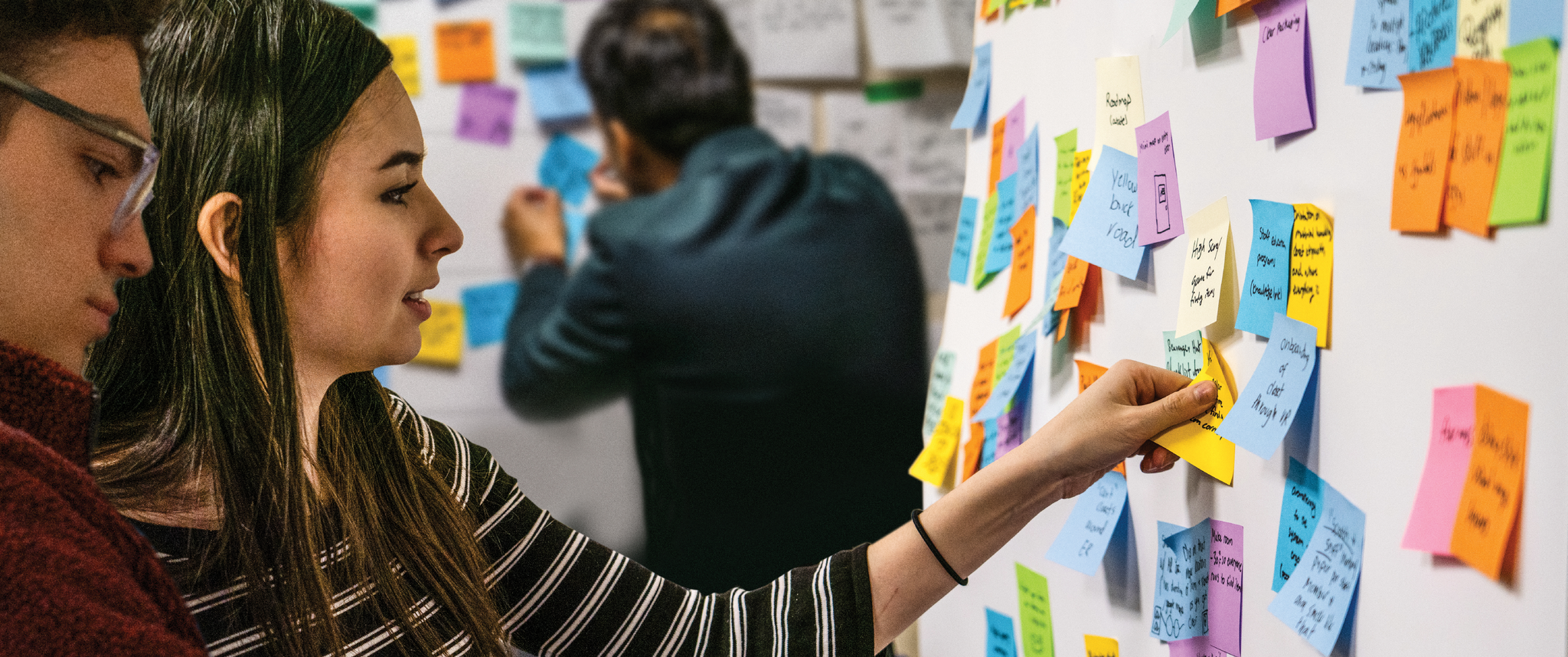 Graduate students place multi-colored sticky notes on two large white boards 