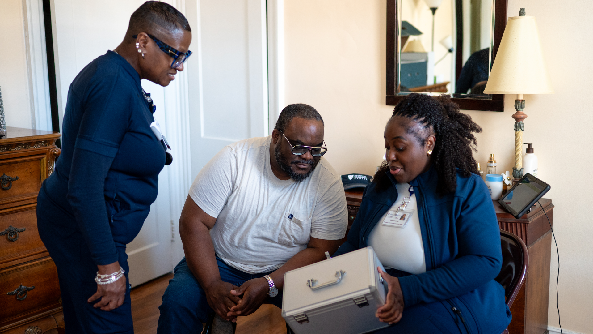 One nurse is standing and another is sitting on either side of James Wright, who is playing the part of patient in the Hospital at Home simulation. One nurse is holding a silver lock box used for storing medications
