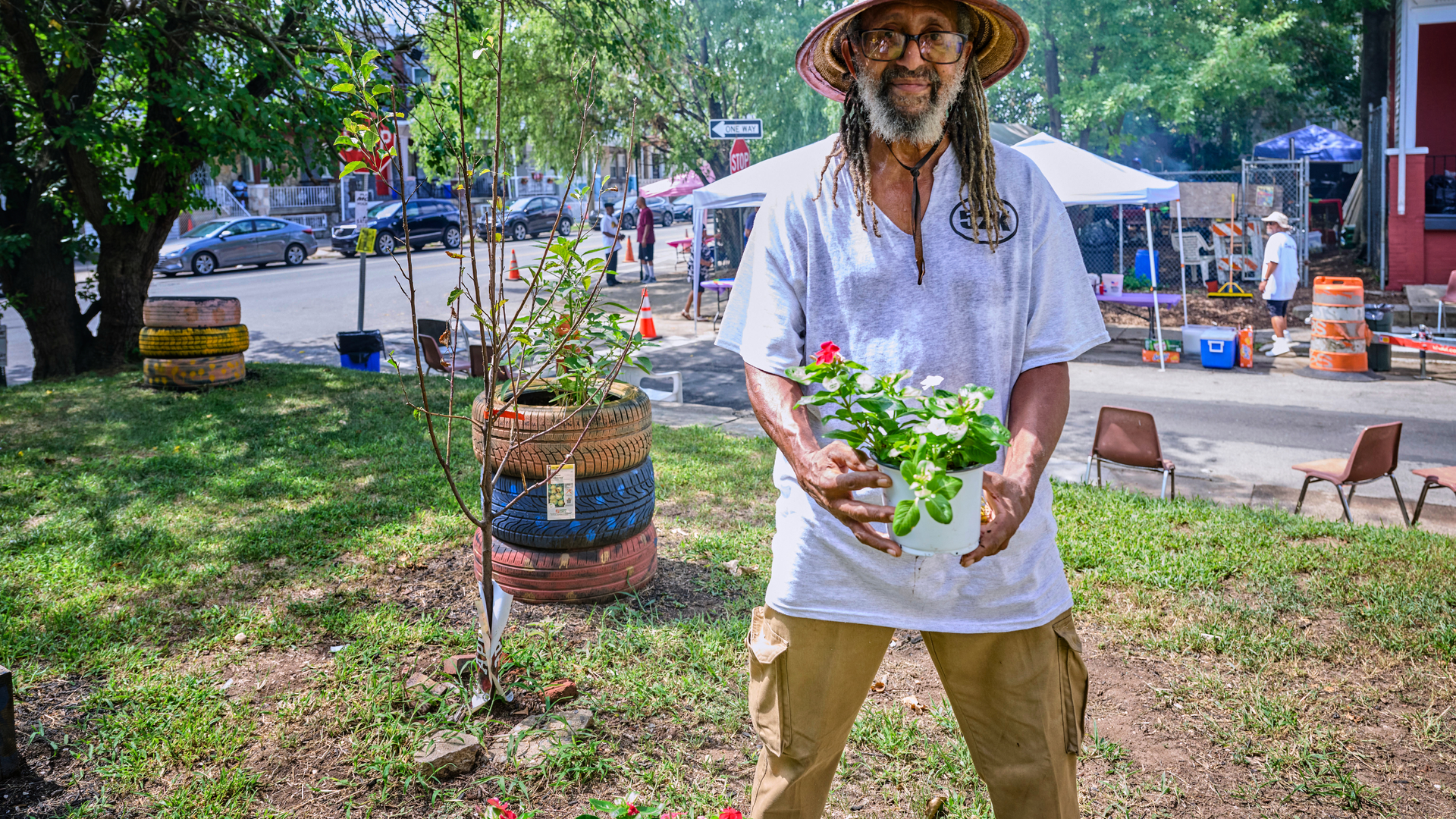 A Black man with long gray dreadlocks holds a potted flower. He is standing in an urban garden with a newly planted tree, with a street party going on behind him