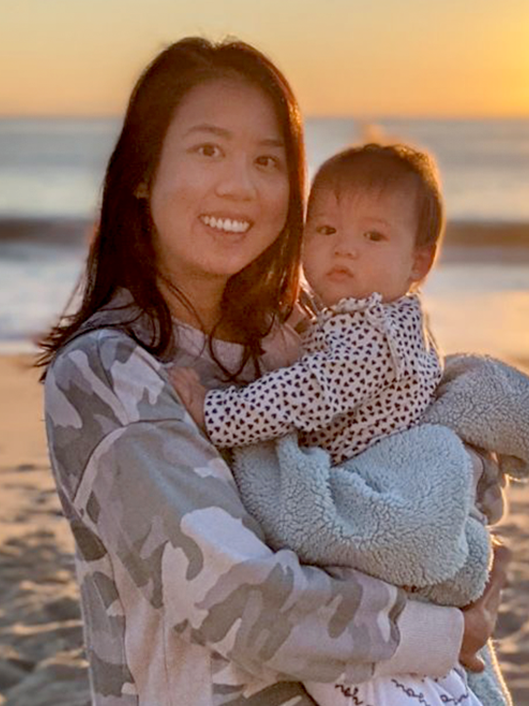 Kristina, a young woman, holds a baby in a blanket on the beach at sunset