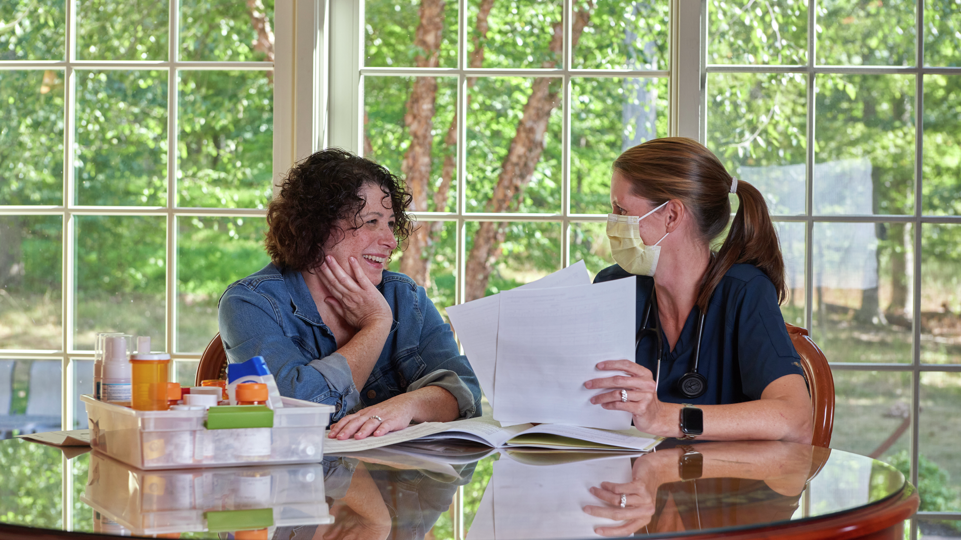 Robin Magaziner, smiling, sits at a dining table with nurse Erin McCarthy, who is showing Robin information in a folder