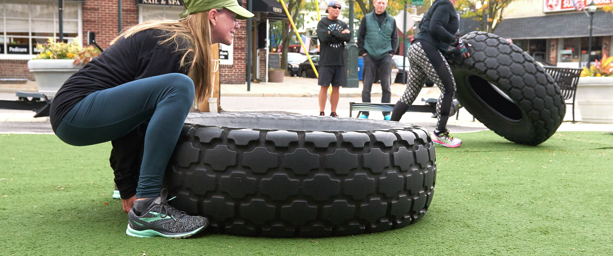Two women flip oversized tires on an urban green as athletic trainers observe
