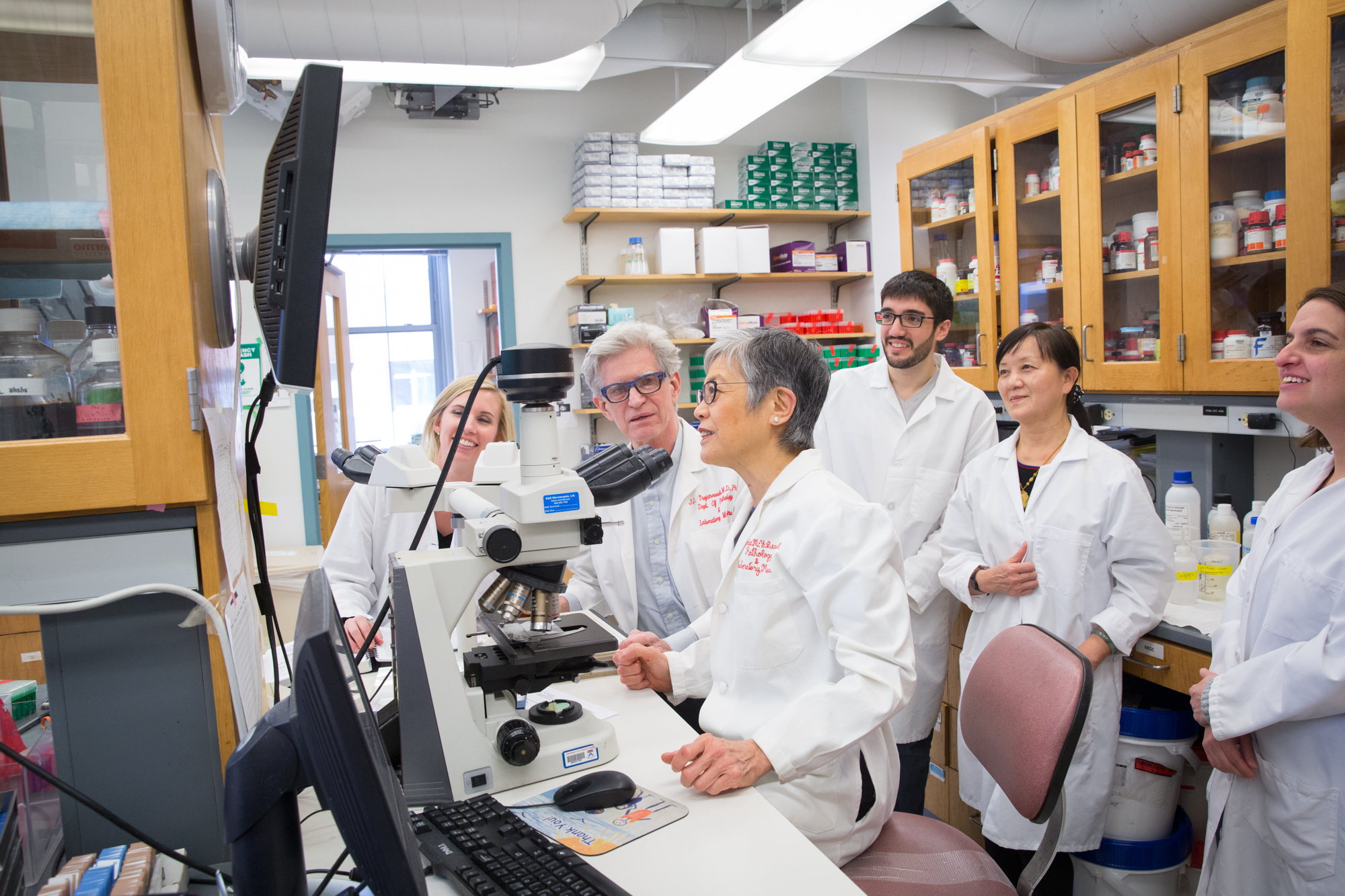 In a lab, John Q. Trojanowski looks toward Virginia Lee, as Lee and a surrounding group of lab members look at a screen in front of her.