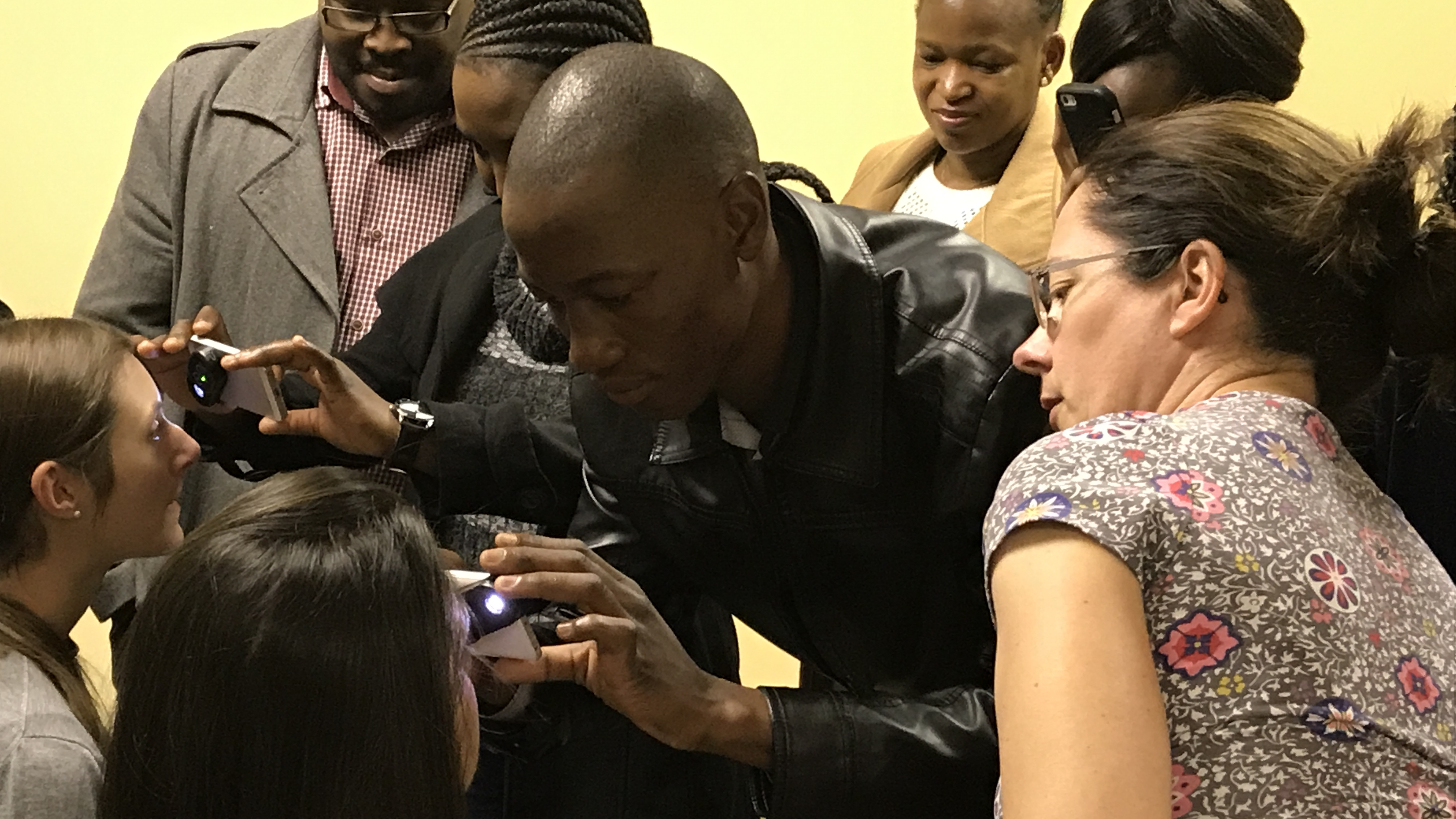 Two people use a device to examine the eyes of seated people, while a group looks on