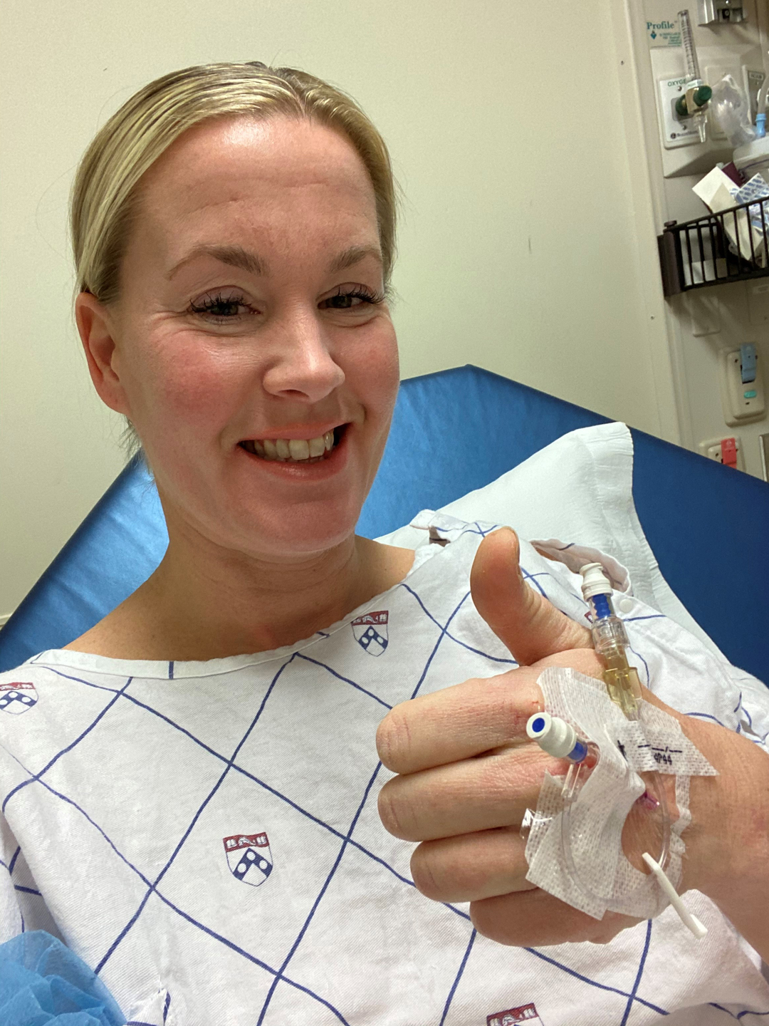 Cheryl Urban, wearing a hospital gown, smiles and gives a thumbs up