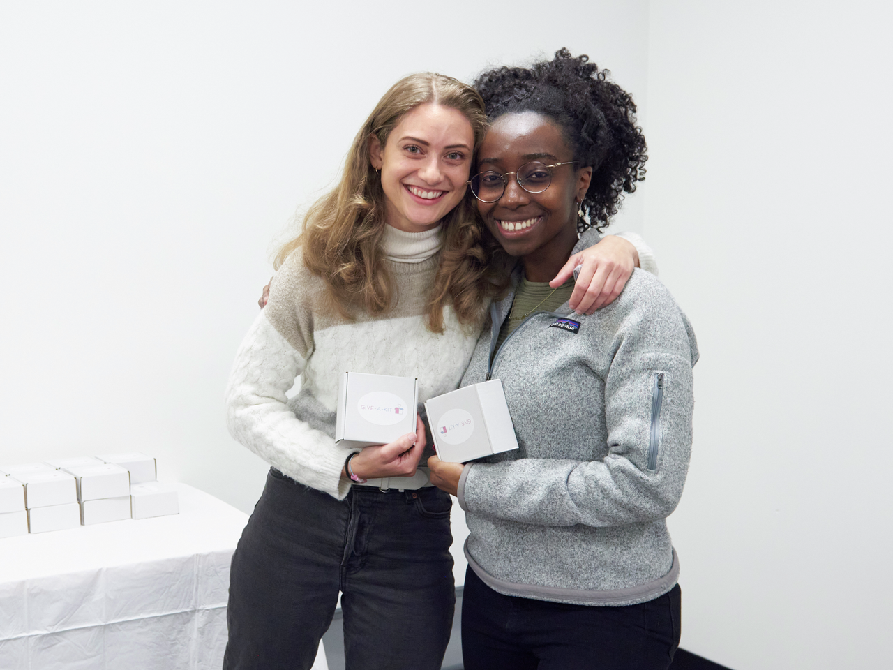 Liz Ramos and Michel Ntiri smile with arms draped on each other's shoulders, holding small boxes marked "Give-A-Kit"