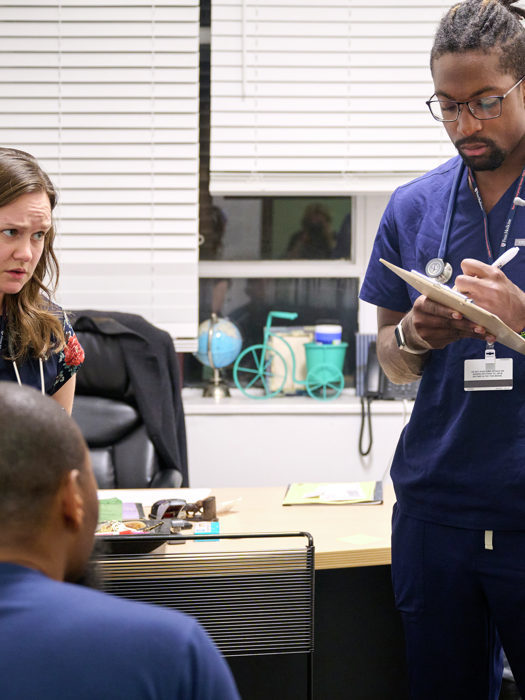 A person in scrubs takes notes on a clipboard, surrounded by other staff and volunteers