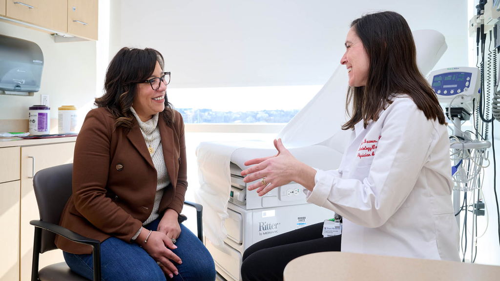Illeana Casiano-Vazquez sits in an exam room with Jessica Heaven. Both are smiling and conversing happily. 
