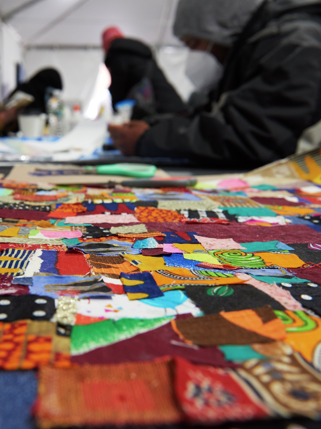 Scraps of colorful fabric overlapping in a quilt are seen in the foreground with figures leaning over the table in the background