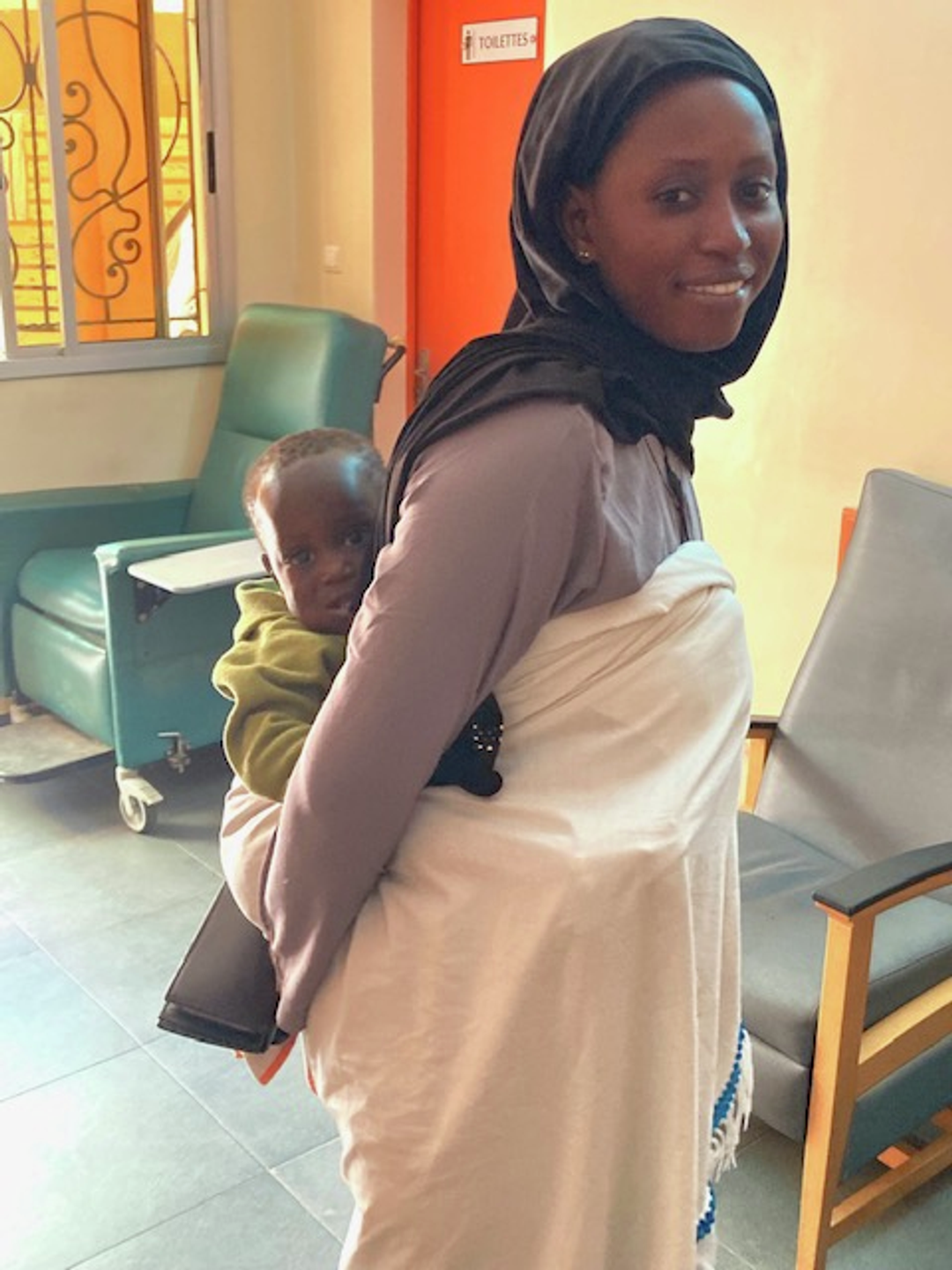 A young woman carrying a child on her back using a cloth wrap in a clinic in Africa. Chairs, a window, and a door labeled 'TOILETTES' are visible in the background.