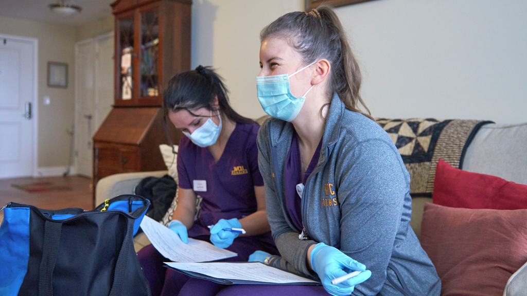 Two student nurses sit on a couch, one taking notes and one engaged in conversation, smiling